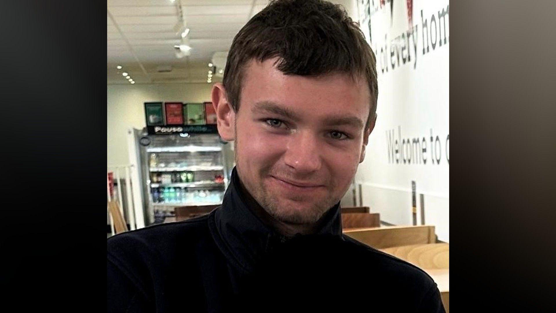 A man wearing a black round-necked top. He has short, brown hair and stubble and he is smiling.