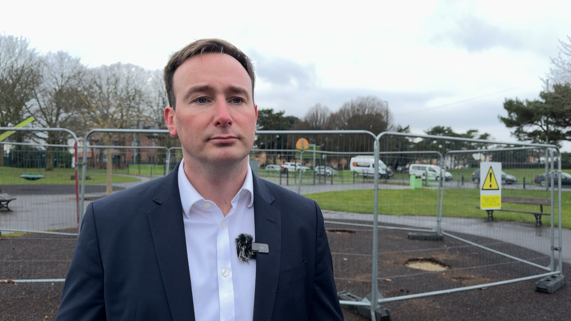 Tom Hayes the MP for Bournemouth East. He is wearing a white shirt and a dark blue jacket. Mr Hayes is standing in a playground in front of a metal fence. Behind you can see a grass area, trees and cars parked on a road.