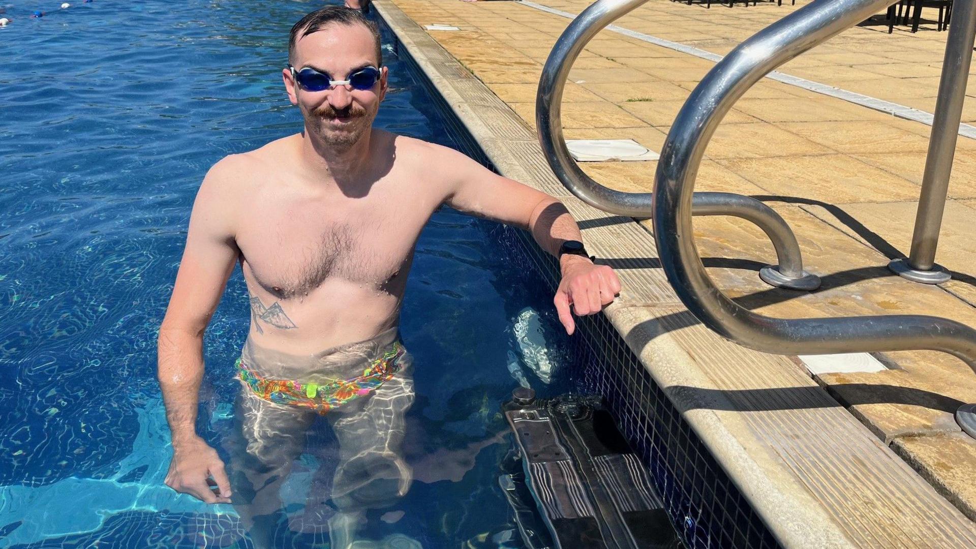 Andy Gilmour-Jones is in the pool wearing goggles. He is posing for the camera with his hand resting on the side of the pool.