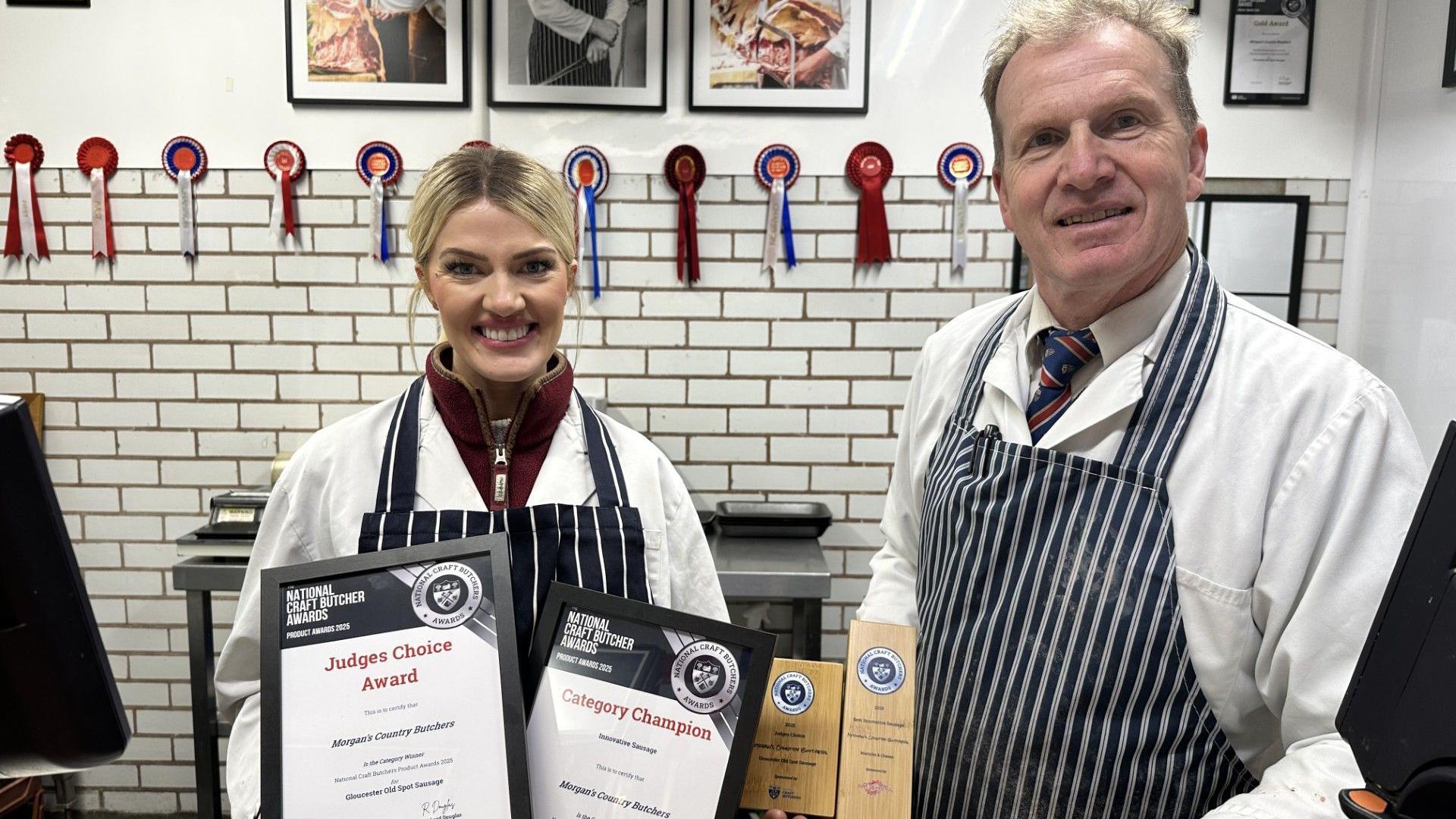 A woman with tied-back blonde hair and a man with short fair hair are both dressed in wite coats and blue-and-white striped butcher's aprons. They are in a white-tiled butchery with rosettes along the top of the tiles and framed product photos above. The pair are holding framed certificates and wooden awards in their hands