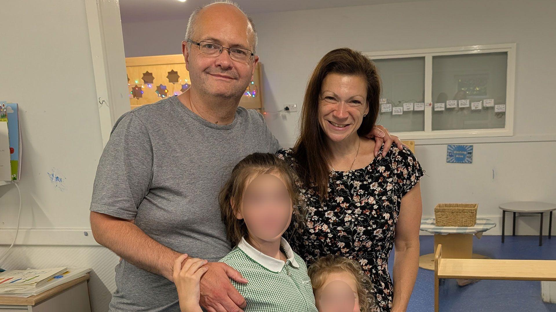 Maxie Allen (right) and his partner Rosalind Levine smile while posing for a photo and looking directly at the camera. Their two children are in the posed family shot but their faces are blurred.