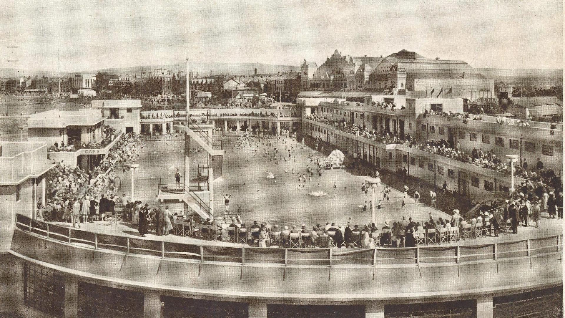 A black-and-white photo shows Morecambe Super Swimming Stadium, with crowds sitting in the grandstands around a large pool.