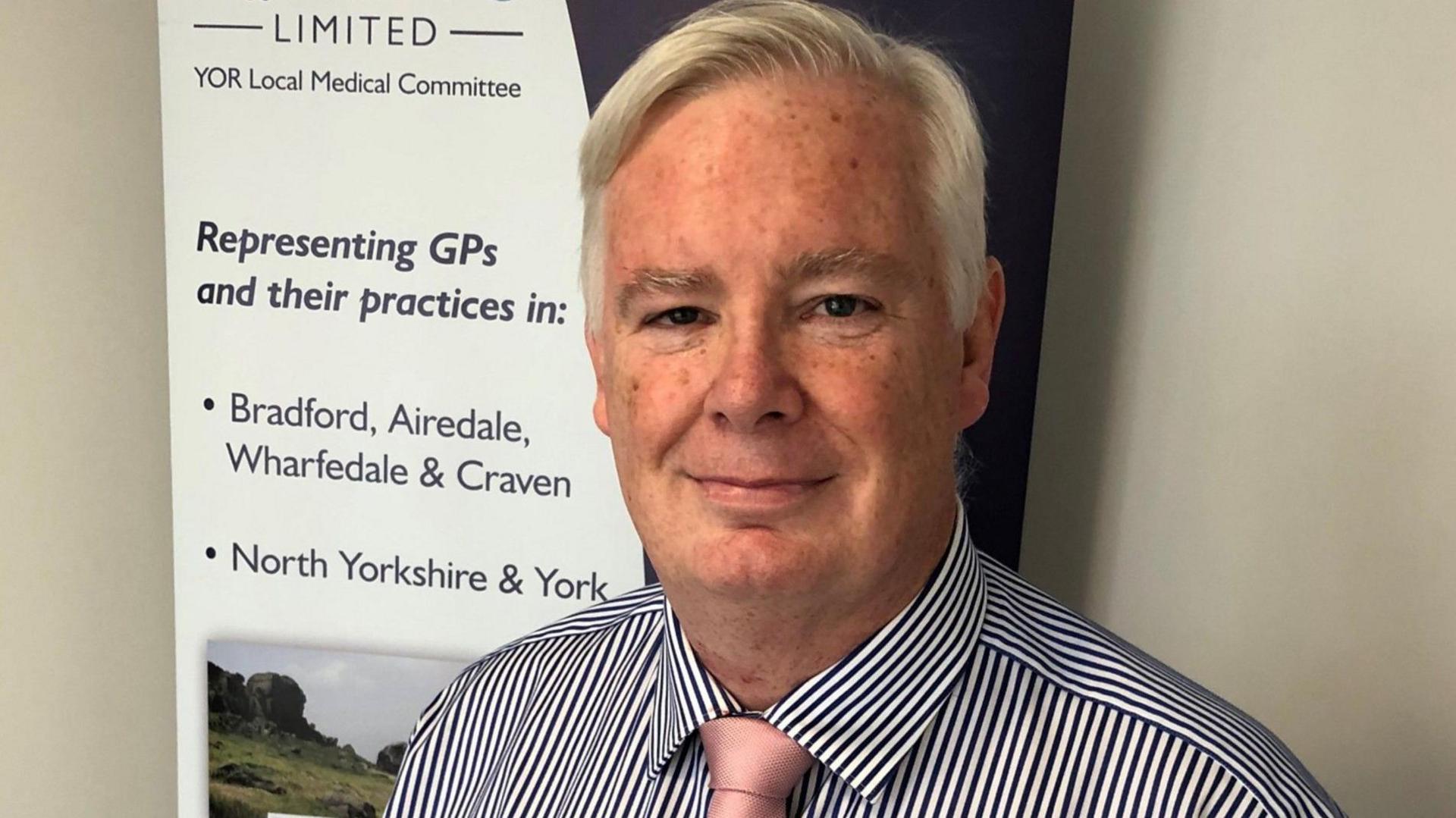 A man stands in front of a YOR Local Medical Committee banner. He has grey hair, and is wearing a pale pink tie and a navy striped shirt. He is smiling.