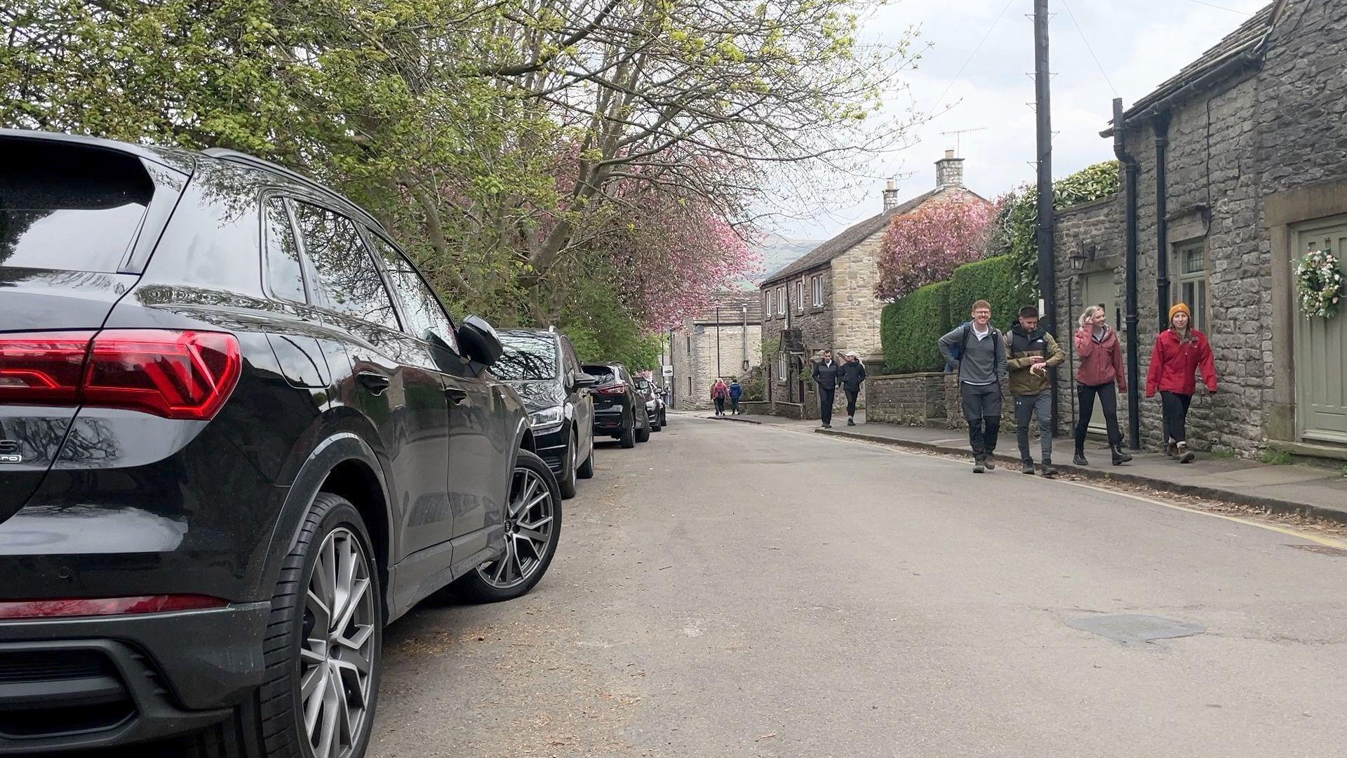 Cars parked in a street in Castleton with a group of hikers walking on a pavement opposite