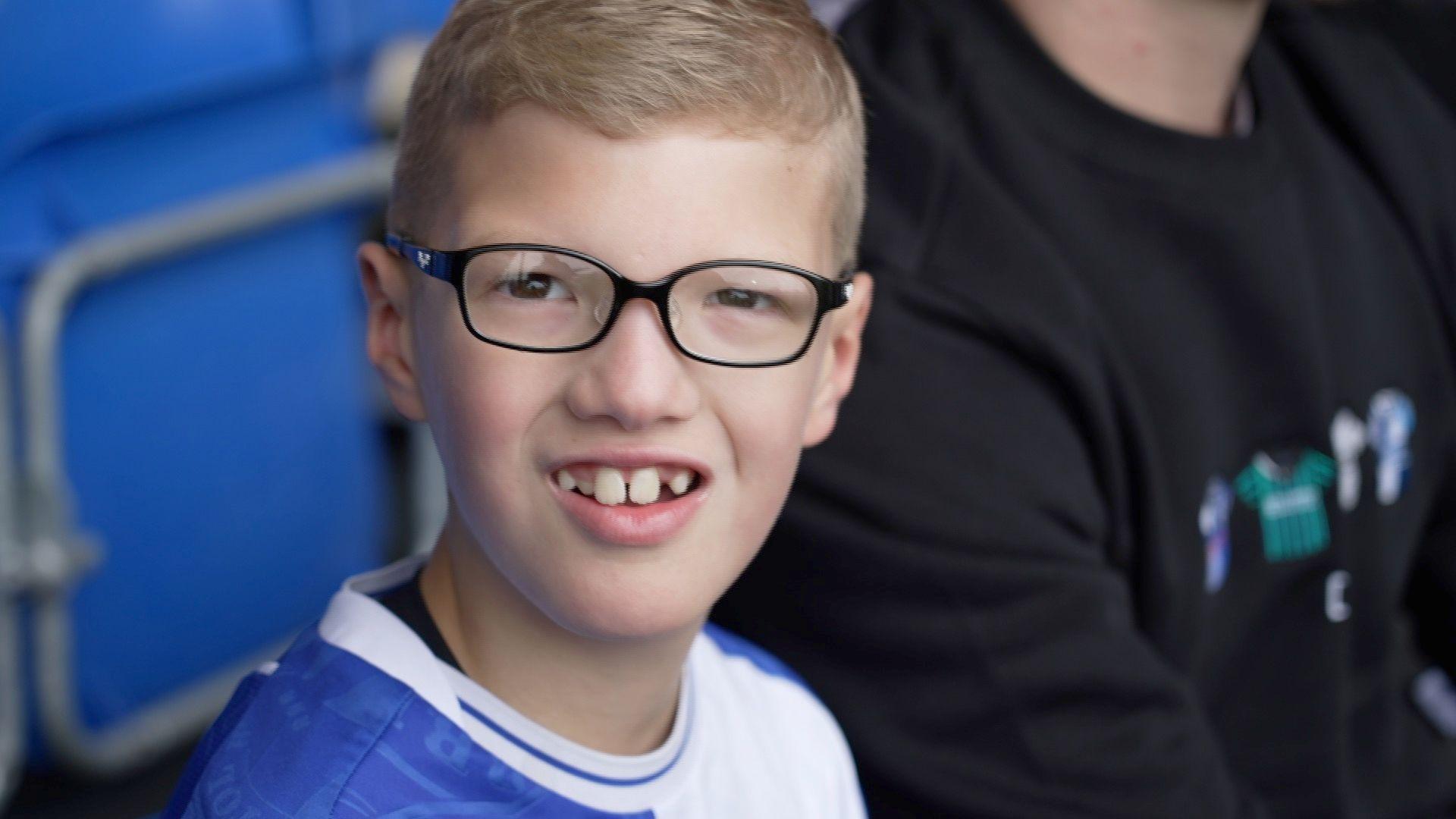 A nine year old boy with glasses looks into the camera. He is wearing a blue and white football shirt.