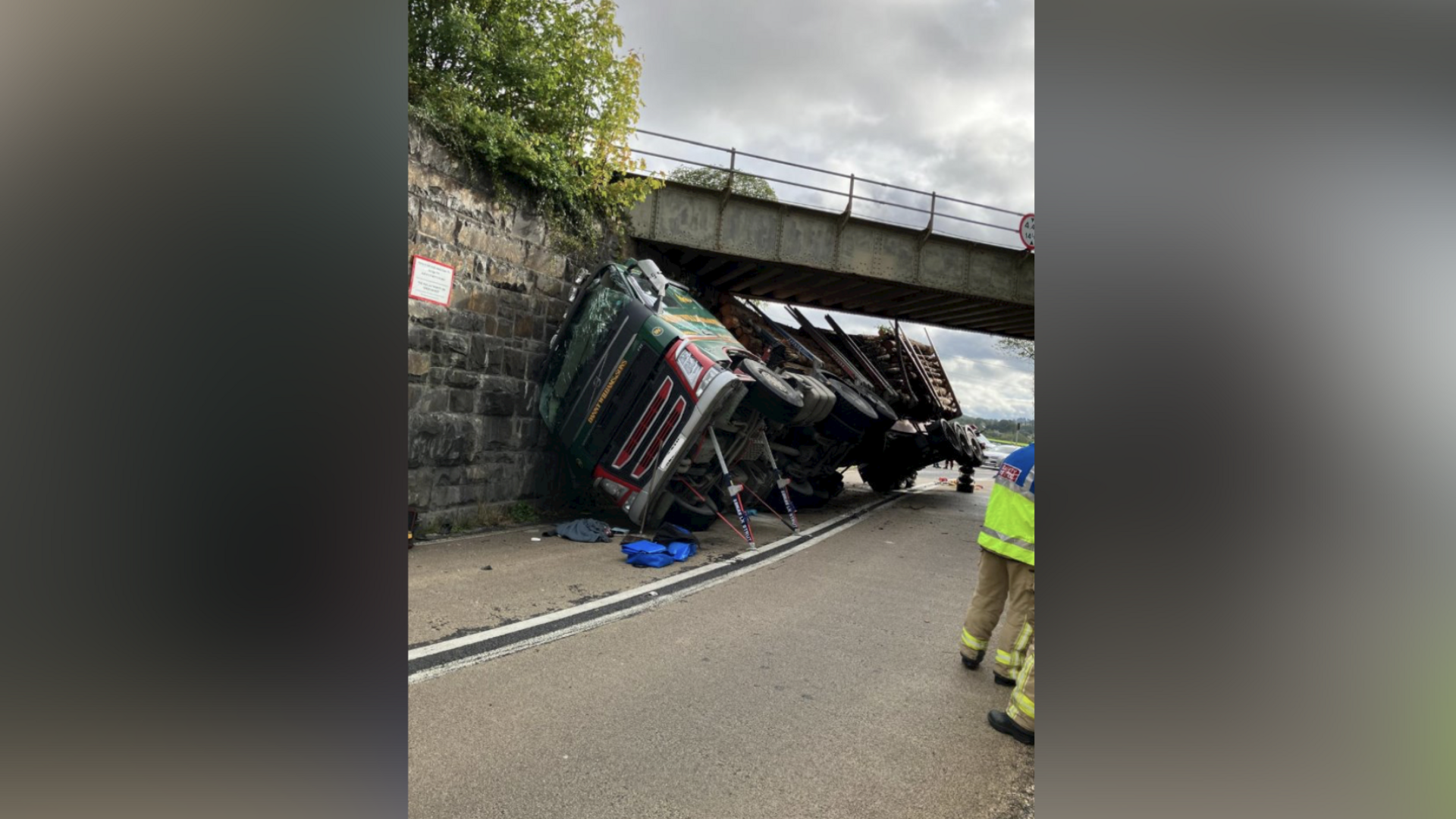 A lorry carrying chopped down trees has overturned and crashed into a railway bridge. It's leaning against the bridge.