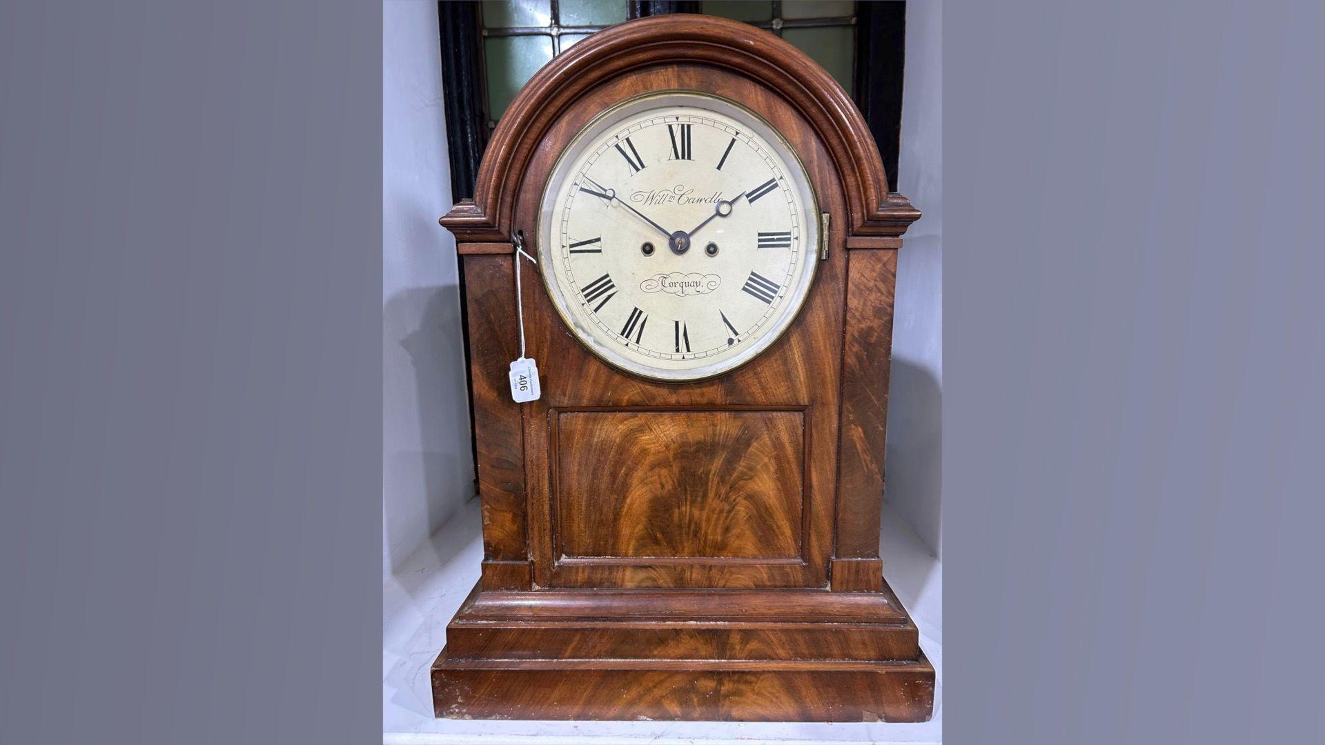 A small brown clock with a white clock face and the numbers in roman numerals. The photo is on a light purple background.