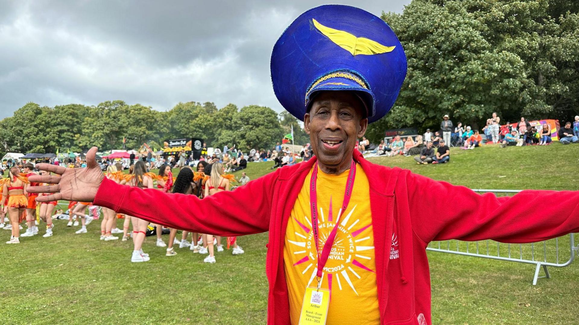 Dr France is wearing a blue headpiece, a yellow t-shirt with the carnival logo in the centre and a red, branded sweater with the zipper open. Around his neck is a carnival pass with his name on it. He has his arms stretched out from side to side and is smiling at the camera.