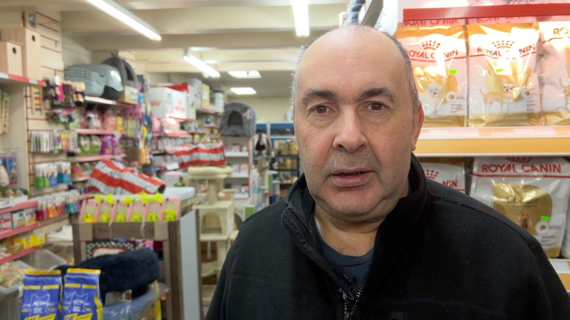 Paul Beresford, wearing a black zip up jumper, looking at the camera. He is stood inside of a corner shop with various food items stacked on shelves behind him. 