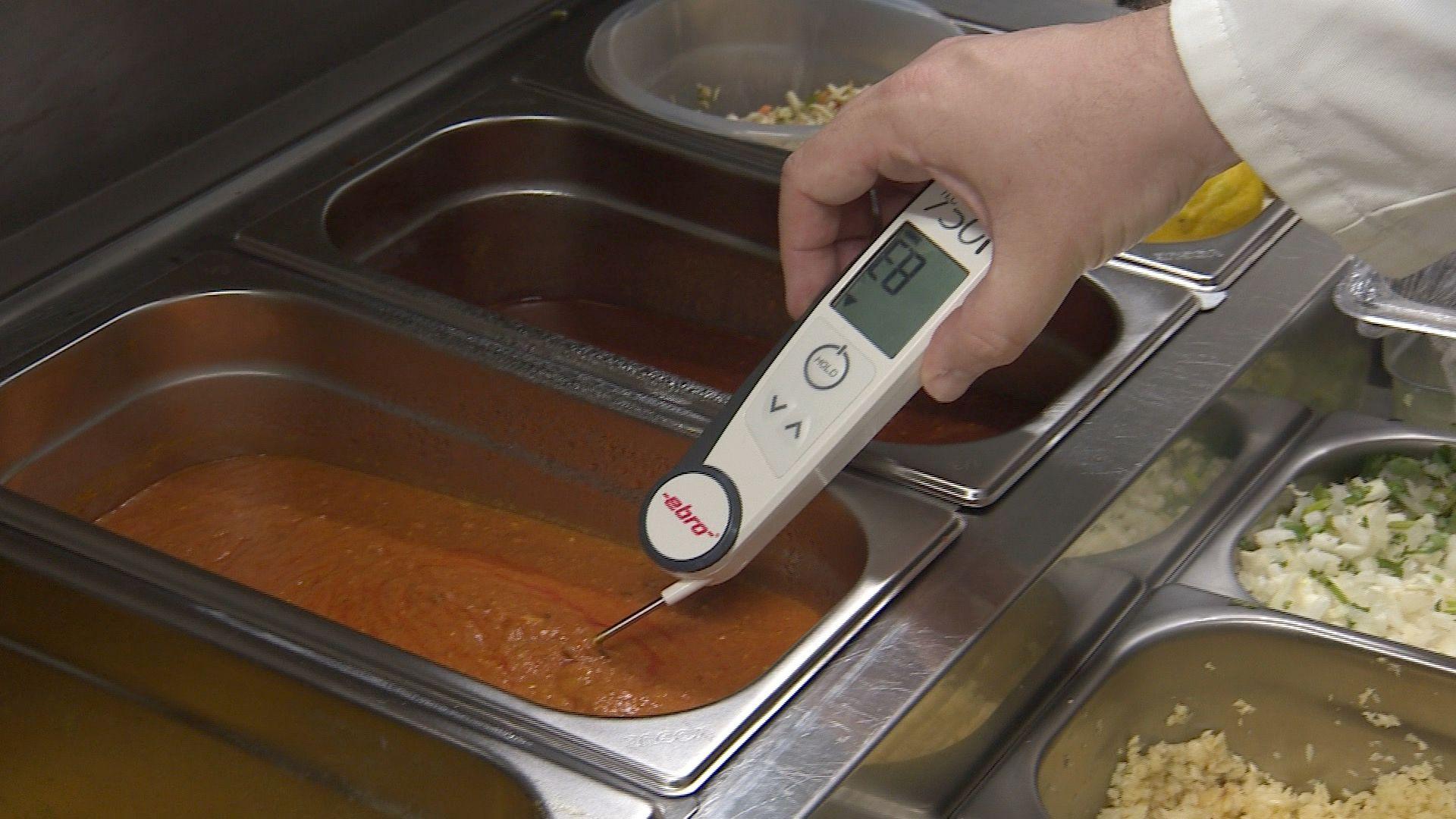 A white food thermometer held by a licensing officer is dipped into a container of curry. The reading on its LCD screen shows it is at 83F.