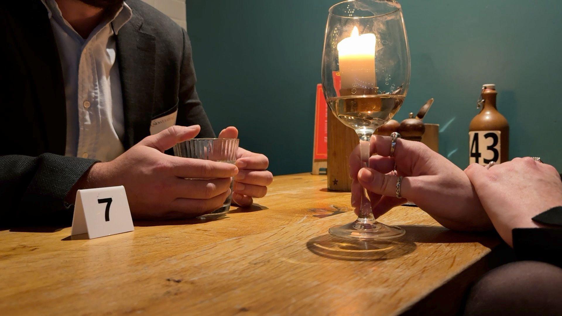 A man and a woman hold their hands against their drinks glasses. There is a number seven on the table. There is a candle on the table as well. The table is brown. 