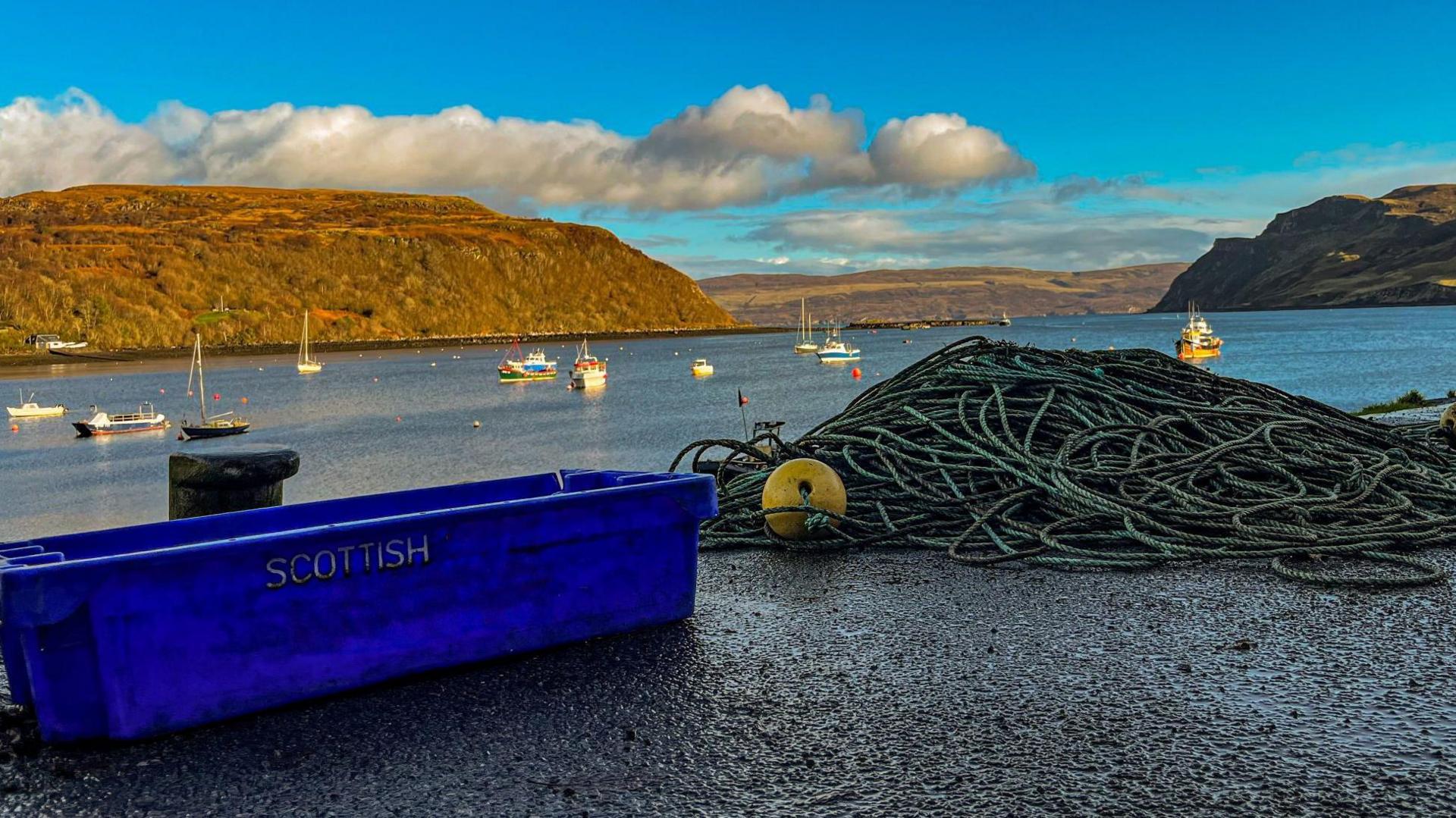An empty blue box with the word Scottish on the side sat next to a pile of ropes on the side of a harbour with fishing boats in the background. 
