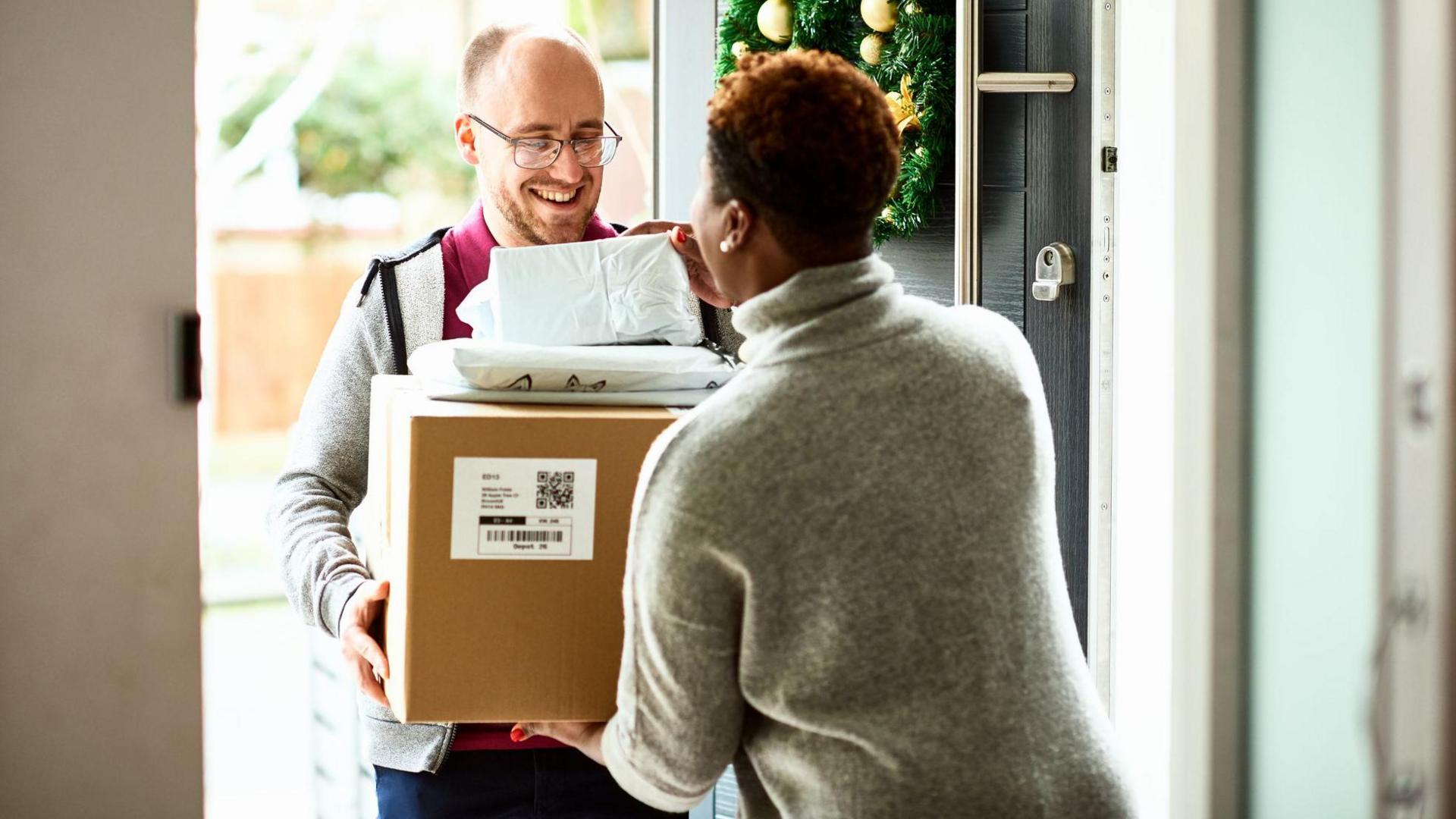 A bald delivery man, wearing a red t shirt and grey zip-up top hands over cardboard box to female customer, at her door. She is only seen from the back, and is wearing a grey jumper and has short, curly dark hair. There is a Christmas wreath on the door.