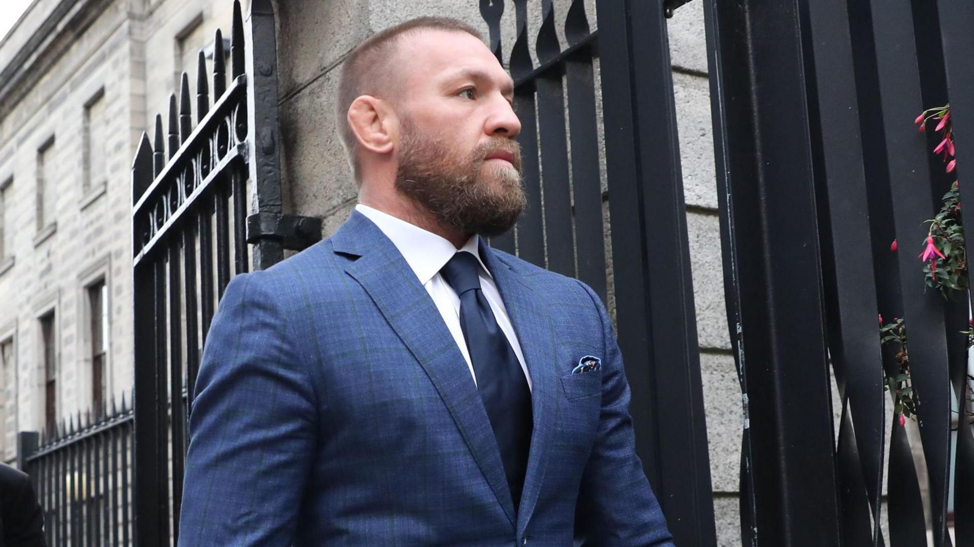 Two men walk out a black gate, one behind the other. The man at the front is wearing a navy suit, navy tie and white shirt, he has ginger short hair and a ginger beard.
The man behind him wears a black suit and tie with a light blue shirt.