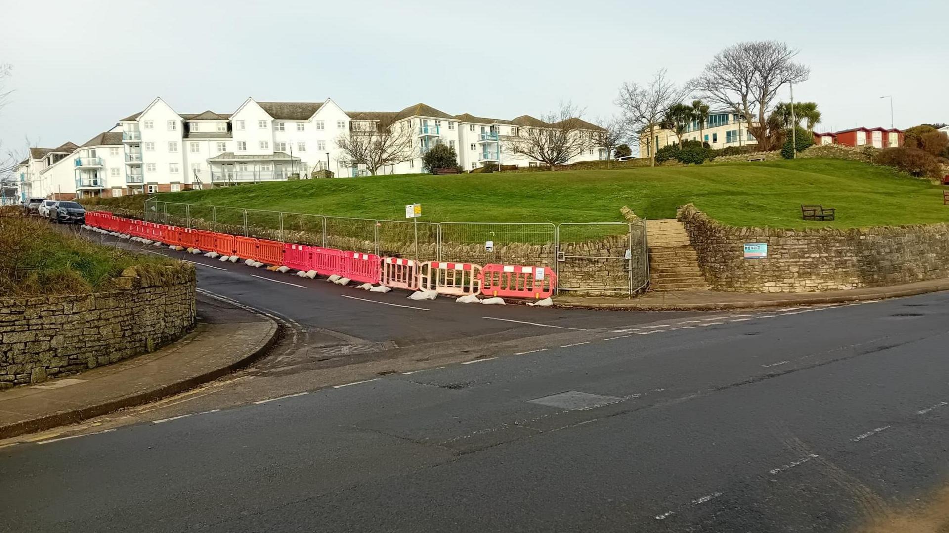 Green area surrounded by a stone wall and road running alongside, red and white barriers are in place along one side of the wall.