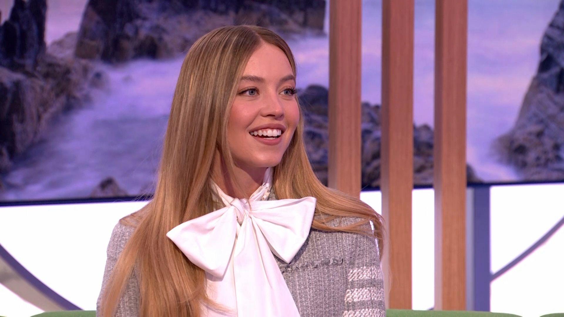 Sydney Sweeney smiles in The One Show studio, wearing a large white bowtie and a grey cardigan. She has long brown hair