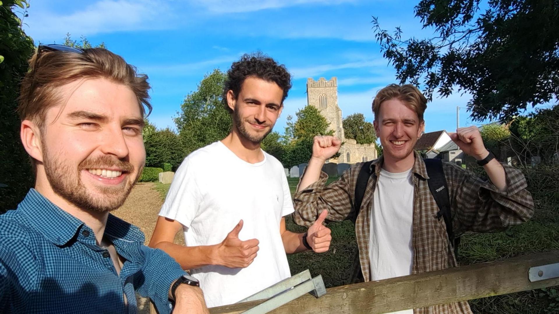 Three men smile in selfie style photo with a church pictured behind them. The man closest the camera has short brown hair with a beard and a blue shirt. The man in the middle has dark curly short hair with a white T-shirt while he gives a thumbs up sign with both hands. The man on the right has raised him arms in triumph and has light short hair with a chequered shirt and white T-shirt underneath.