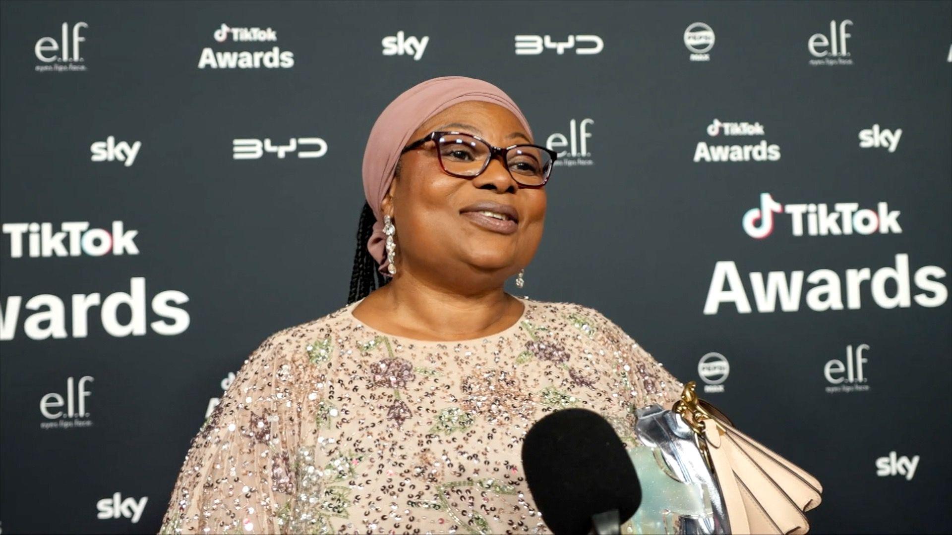 Bemi wears a sparkling beige dress with sequins stands in front of a TikTok Awards backdrop, with a microphone.