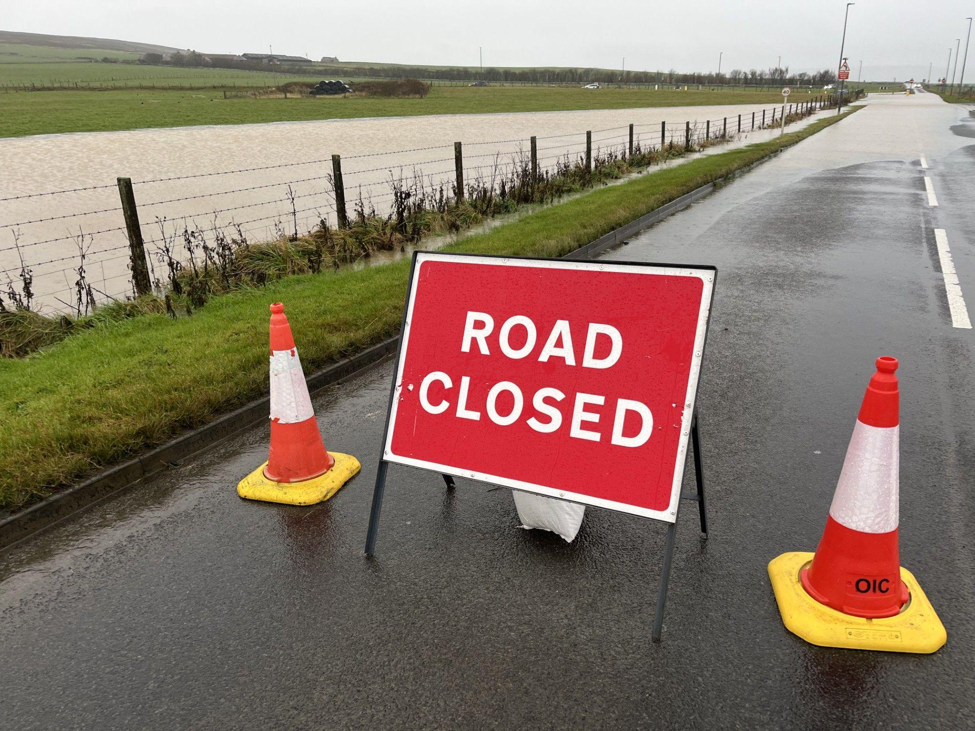 A red road closed sign with two traffic cones either side, sitting on a road and a big pool of water behind.