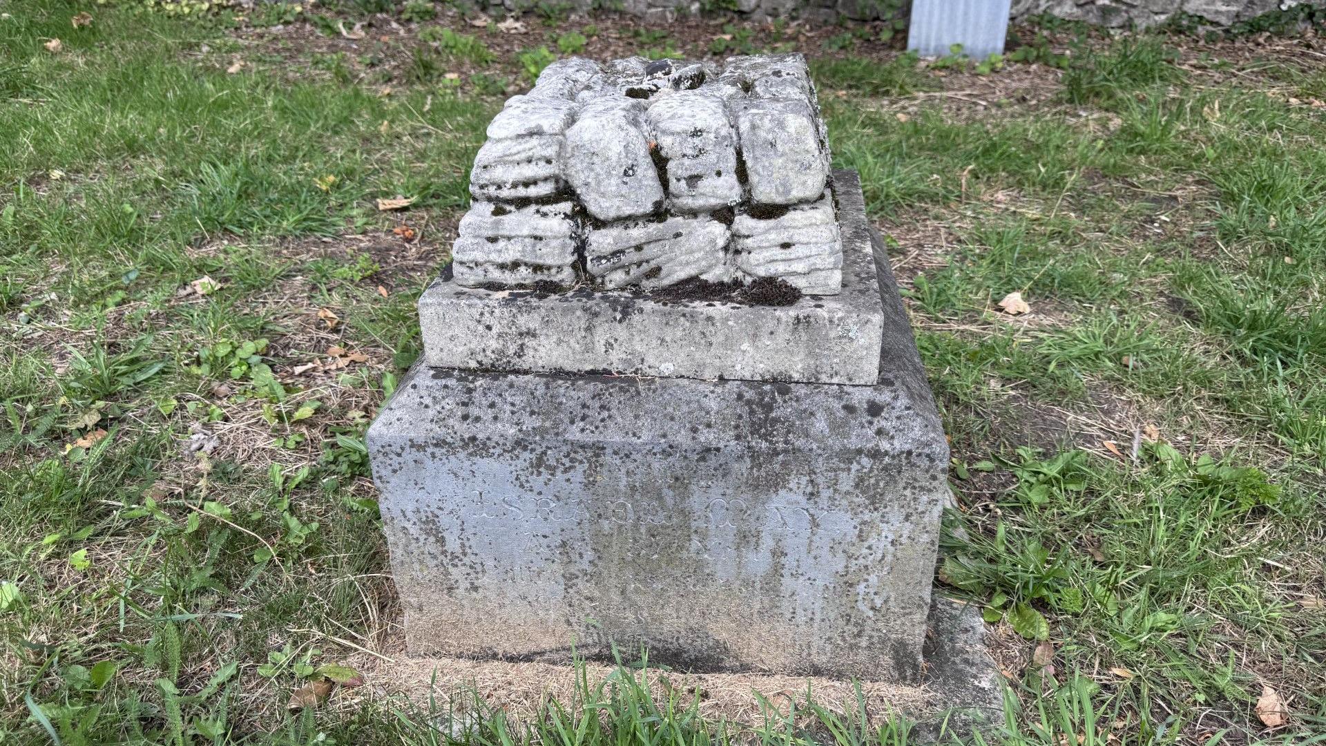 A grave with a number of broken stones on top of it. The grave is dirty and has grass surrounding it.
