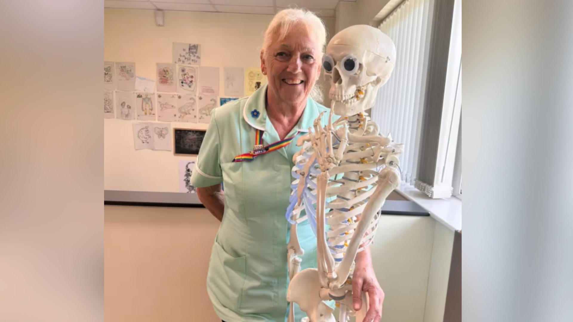 Sue Baker, who has been a nurse for 50 years, pictured beside a skeleton in green scrubs. Sue has white hair and is wearing a rainbow lanyard.