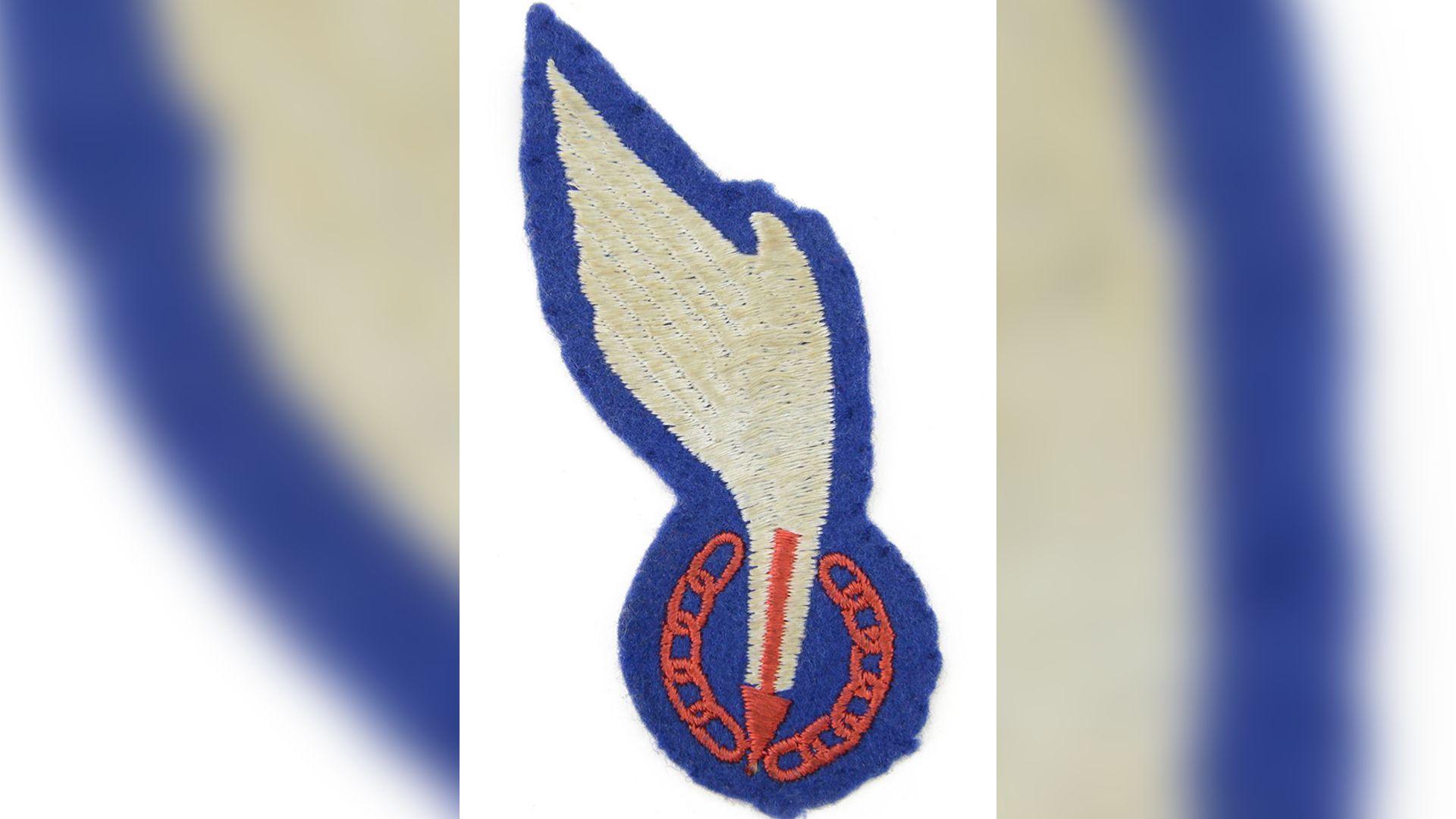 A wing badge of the Special Allied Airborne Reconnaissance Force (SAARF). The embroidered item is a white wing surrounded by blue, with a red arrow and a red chain at the bottom.