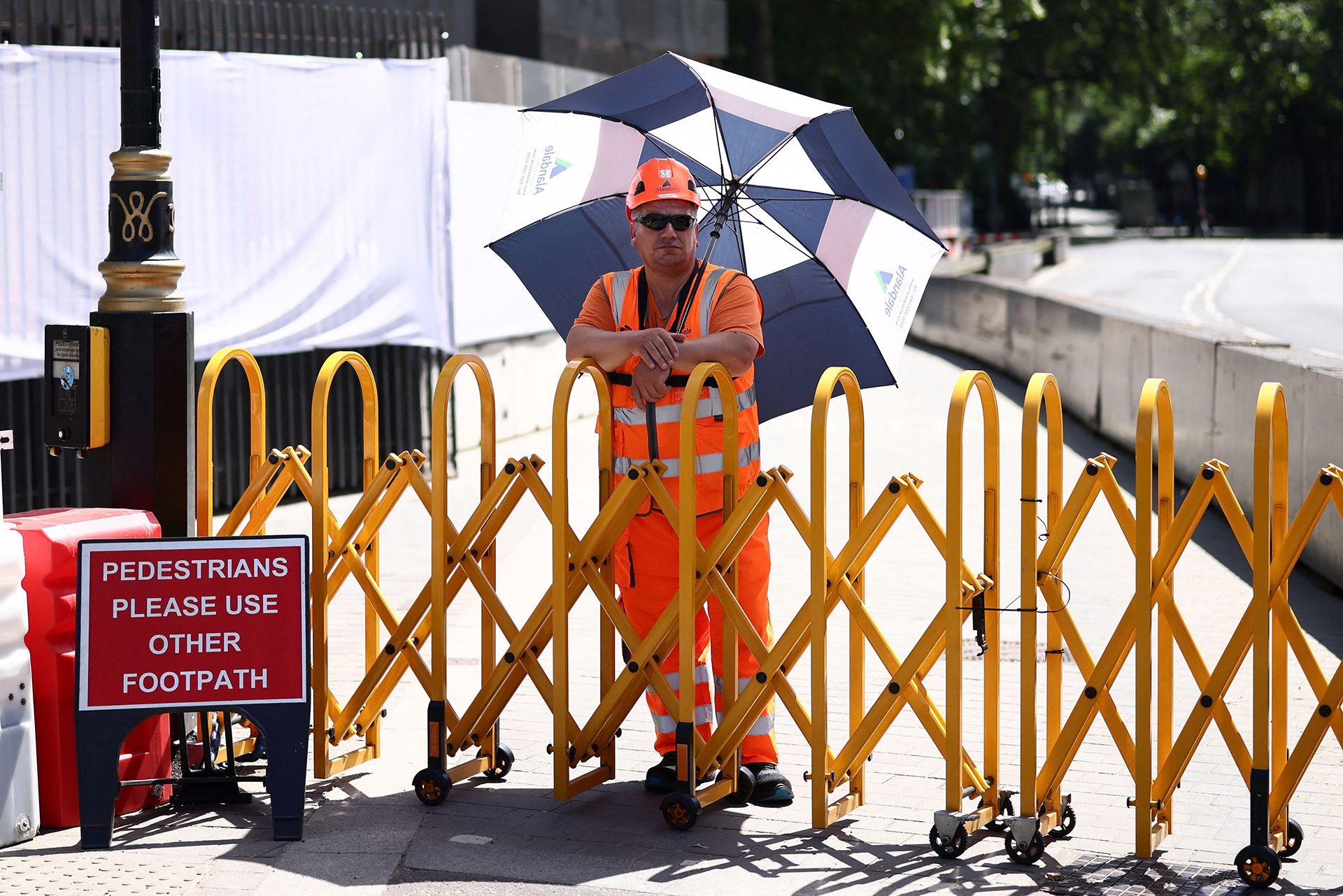 A worker in a high vis orange vest and helmet stands in the shade with a parasol above their head while standing next to a yellow barrier on a closed footpath in London on Tuesday - there is a sign next to him reading 'pedestrians please use other footpath'