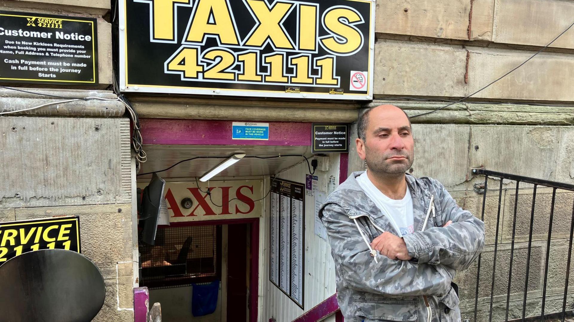 A standing man with his arms folded stood outside building with a sign saying 'Taxis 421111'. He has short, balding hair and is wearing a light-coloured catalogue-style jacket and white T-shirt.