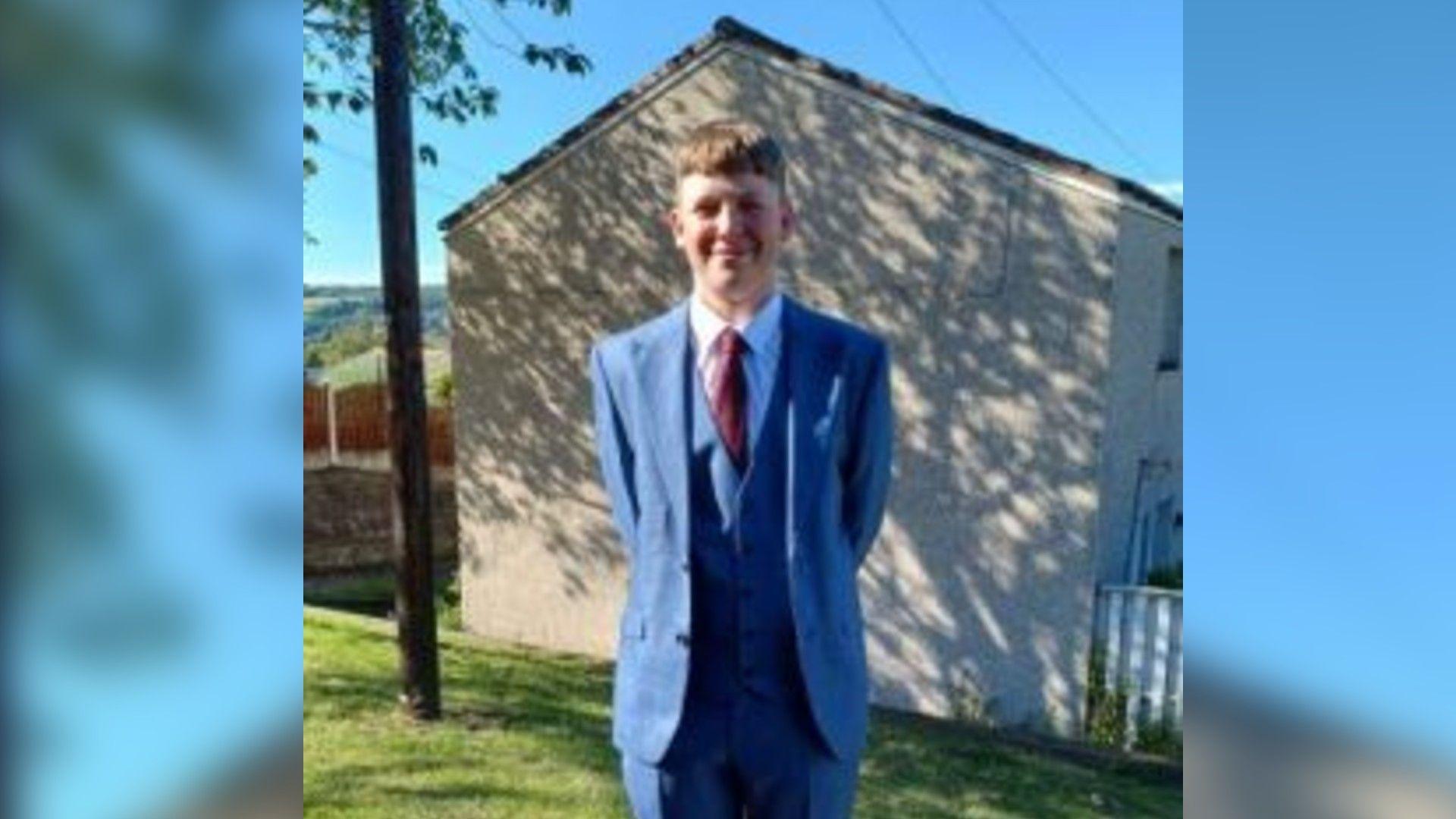 A teenager in a blue suit and red tie stands in a garden on a sunny day
