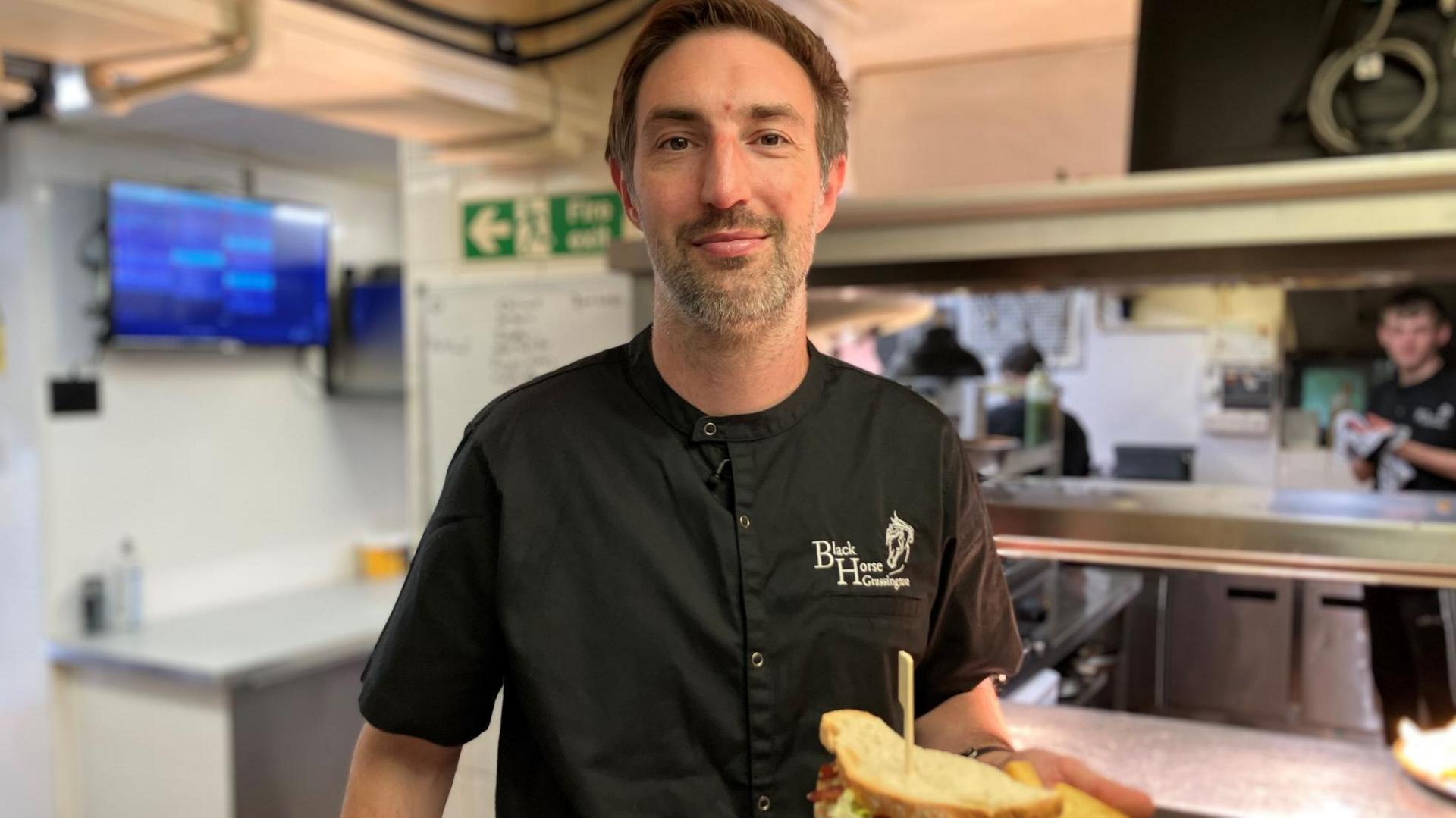 A chef in black uniform holds up a sandwich in a pub kitchen.