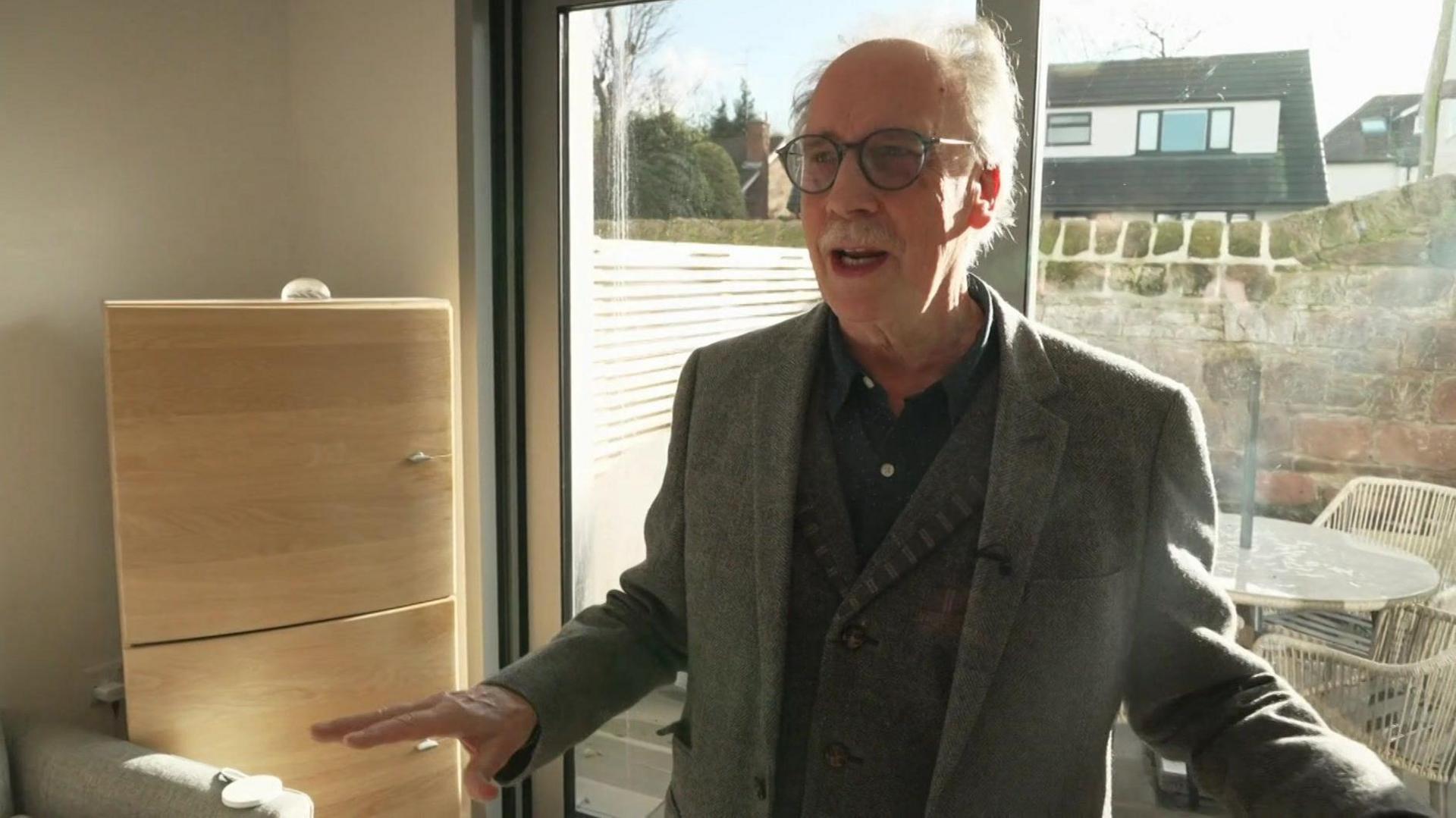 Colin Usher, who has round black-rimmed glasses, white hair and a white moustache, speaks to the camera in front of patio doors looking on to a rear yard