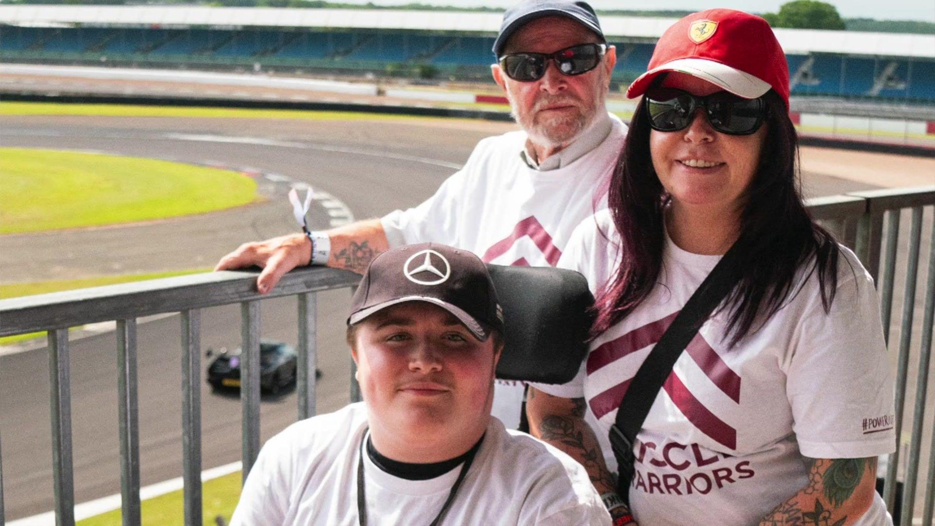 Dakota and his mother and grandfather at the side of the racetrack looking towards the camera. All are wearing caps with car manufacturers on them.