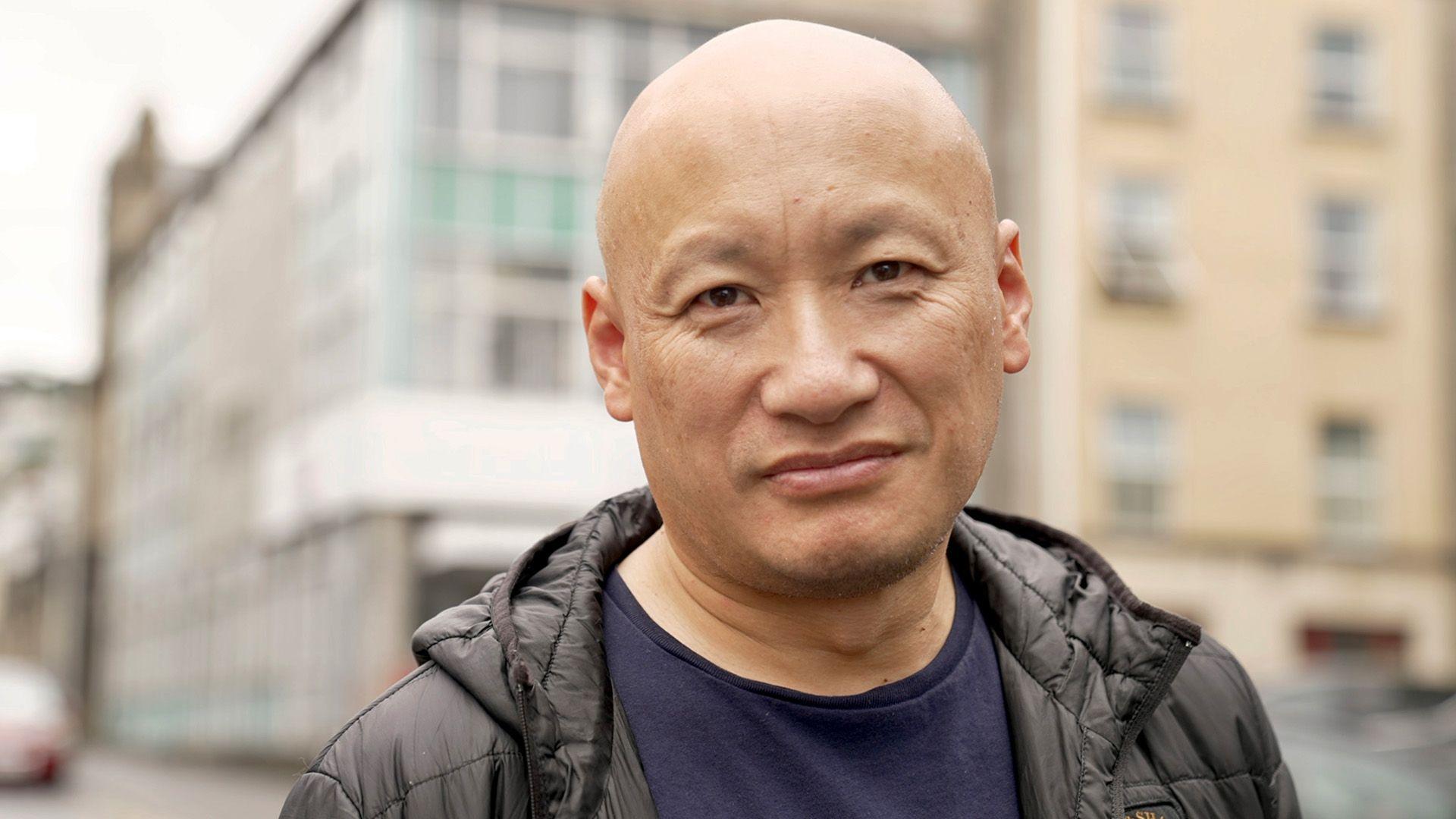 Daniel is standing outdoors, wearing a black jacket over a dark blue shirt. The background features an urban setting with buildings and a street, slightly out of focus."