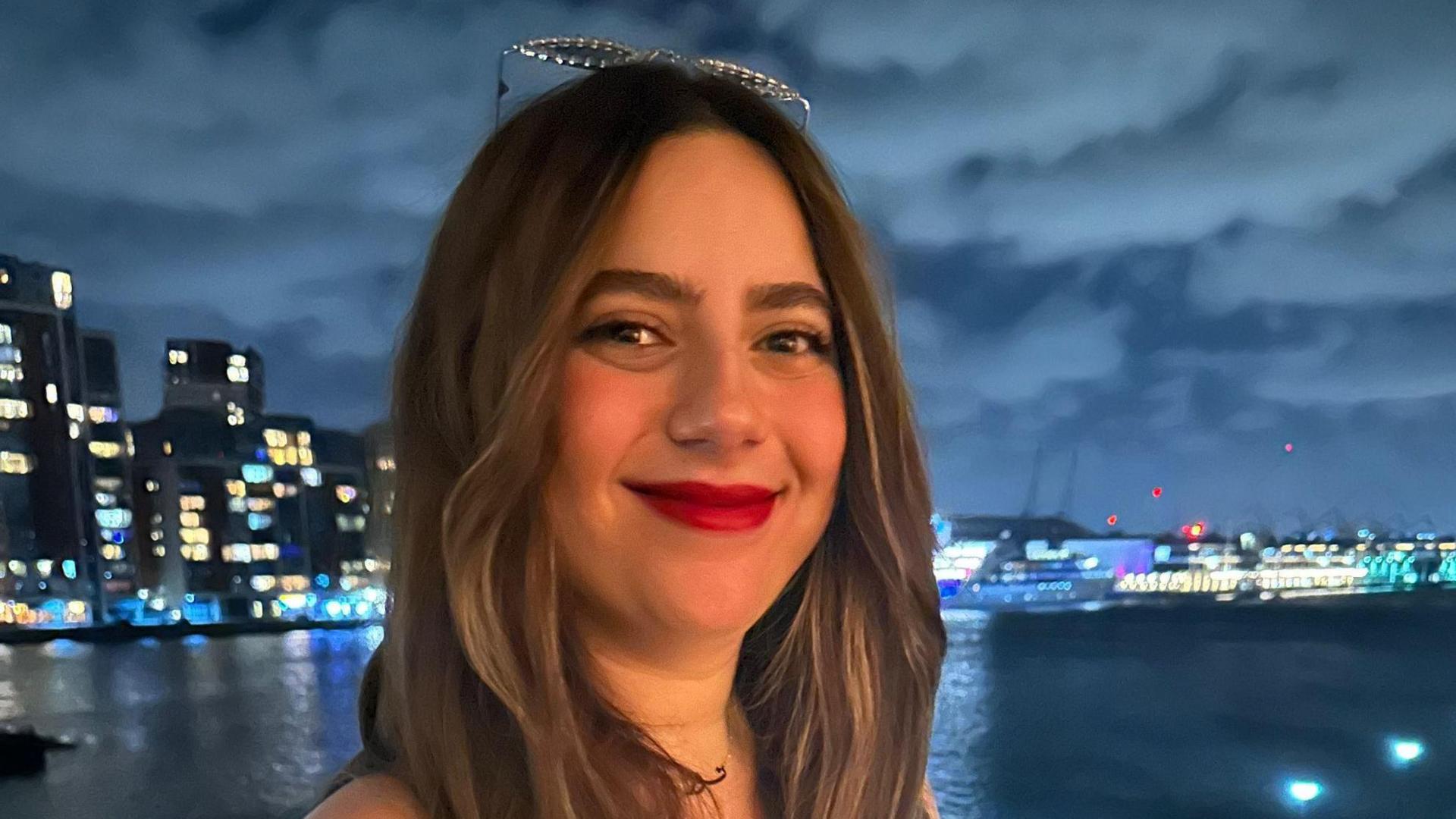 Nura Newman has brown hair and sunglasses on top of her head. She is smiling in front of a wide river at night. Large buildings and boats are illuminated.