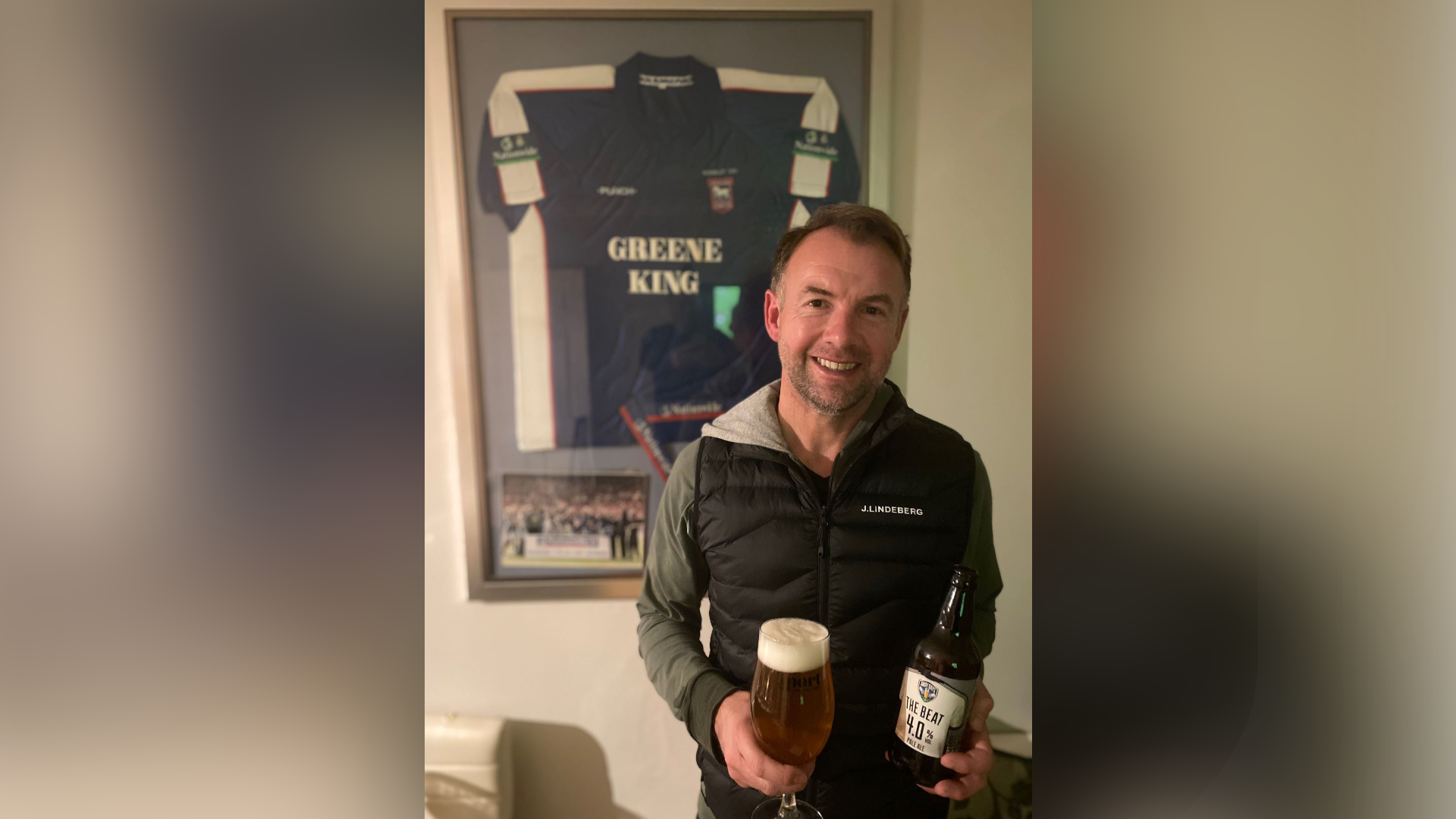 Marcus Stewart is pictured smiling at the camera while holding a beer bottle in one hand and a glass filled with the beer in another. He is wearing a black gilet jacket with a green hoodie underneath. An old Ipswich Town football shirt in blue and white is framed on the wall behind him.