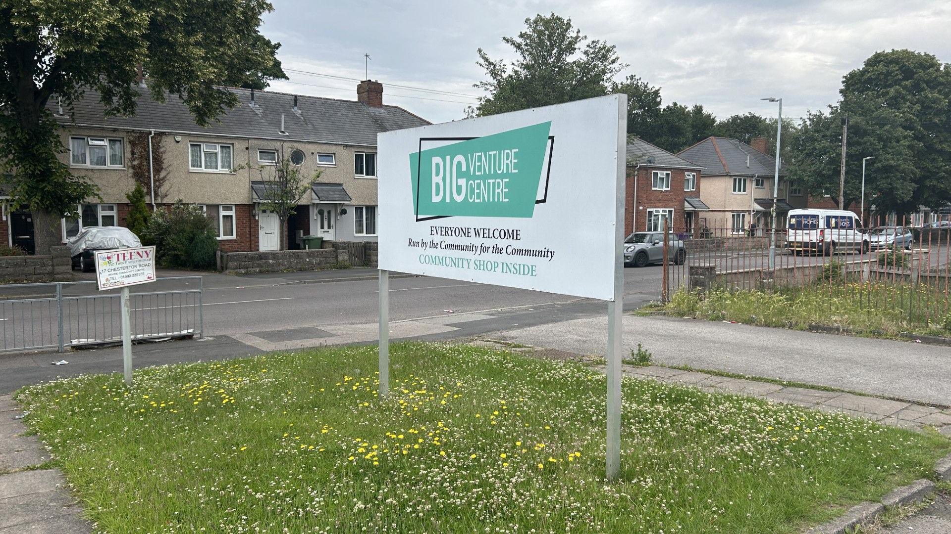 A sign for the Big Venture Centre. There is a grassy area behind with daffodils. We can also see a residential road with some houses in the background.