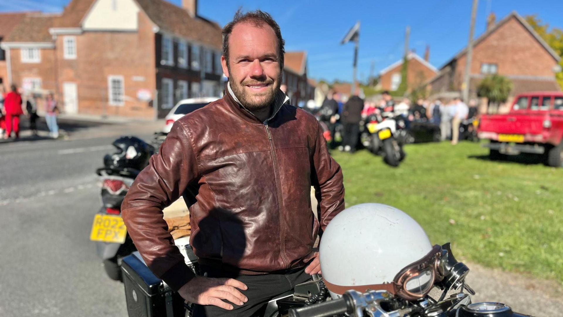 A man wearing a brown jacket while sitting on his motorbike. His hands are on his hips and he is smiling at the camera. It is a sunny day and there is a green or common area behind him.