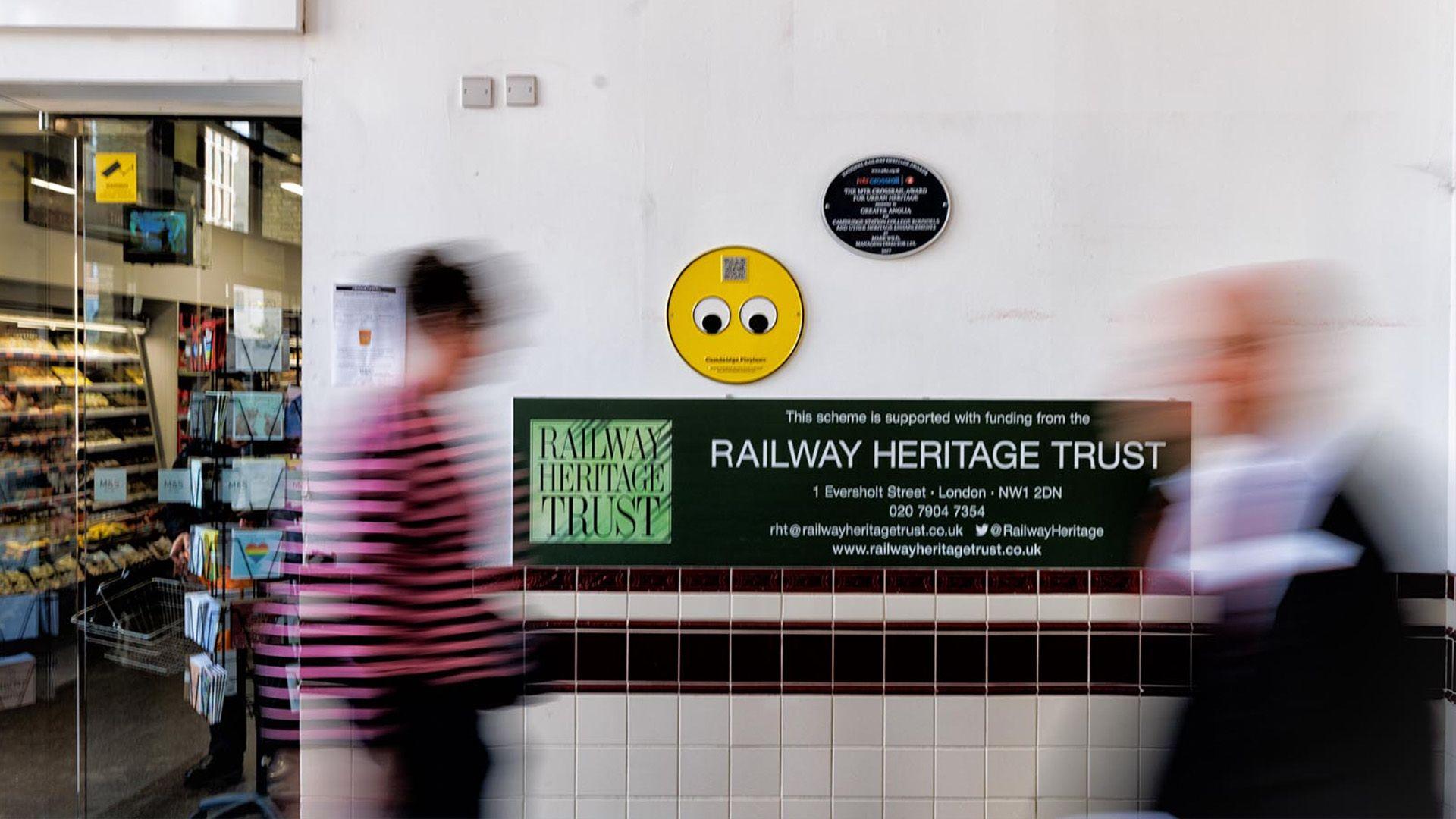 Googly eyes plaque which is just two eyes on a yellow plaque and it's up above a sign for the Railway Heritage Trust while people speed past it.