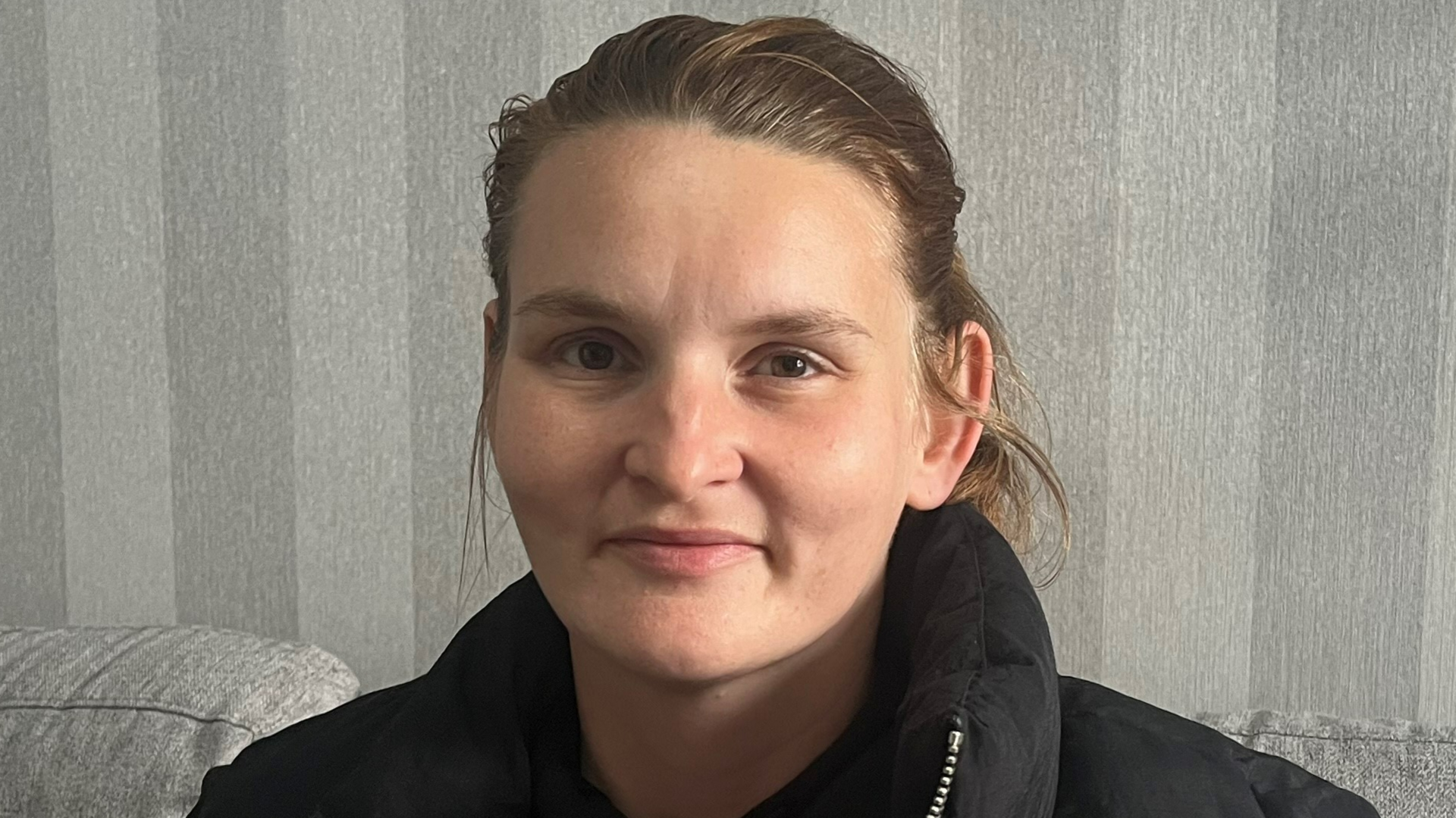 A woman with brown hair tied back wearing a black coat, directly facing the camera, with striped grey wallpaper in the background