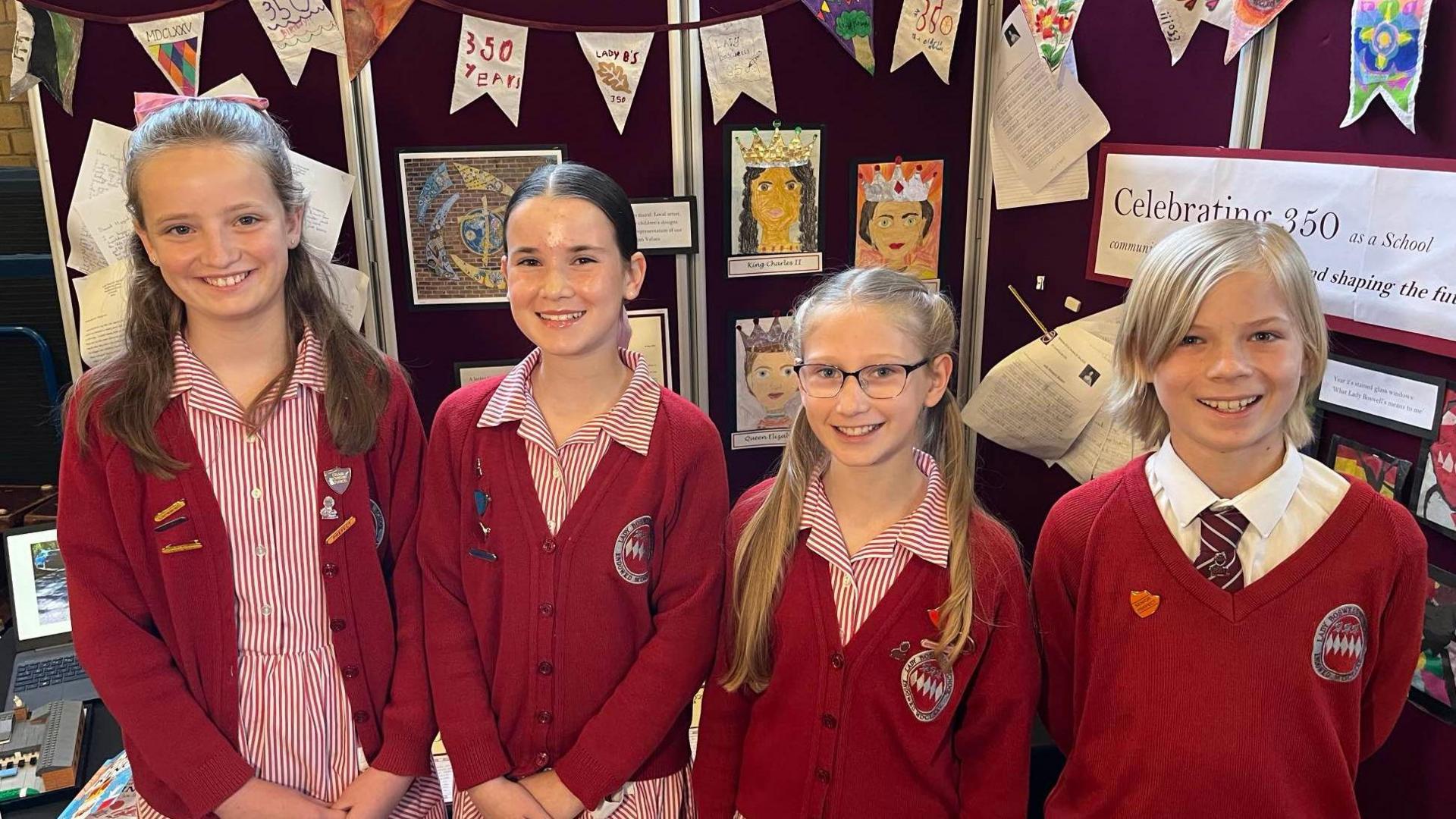 Four primary school children stood side-by-side, all smiling at the camera. They are wearing red card