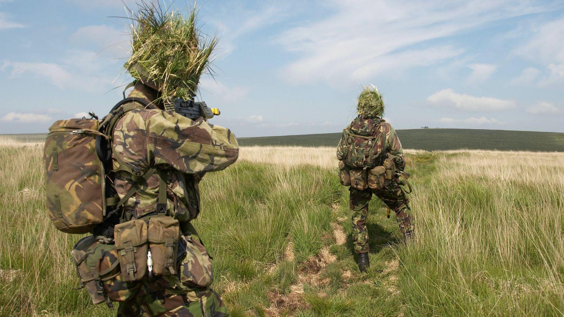 Two camouflaged British soldiers make their way through an open field towards a helicopter on the horizon.  