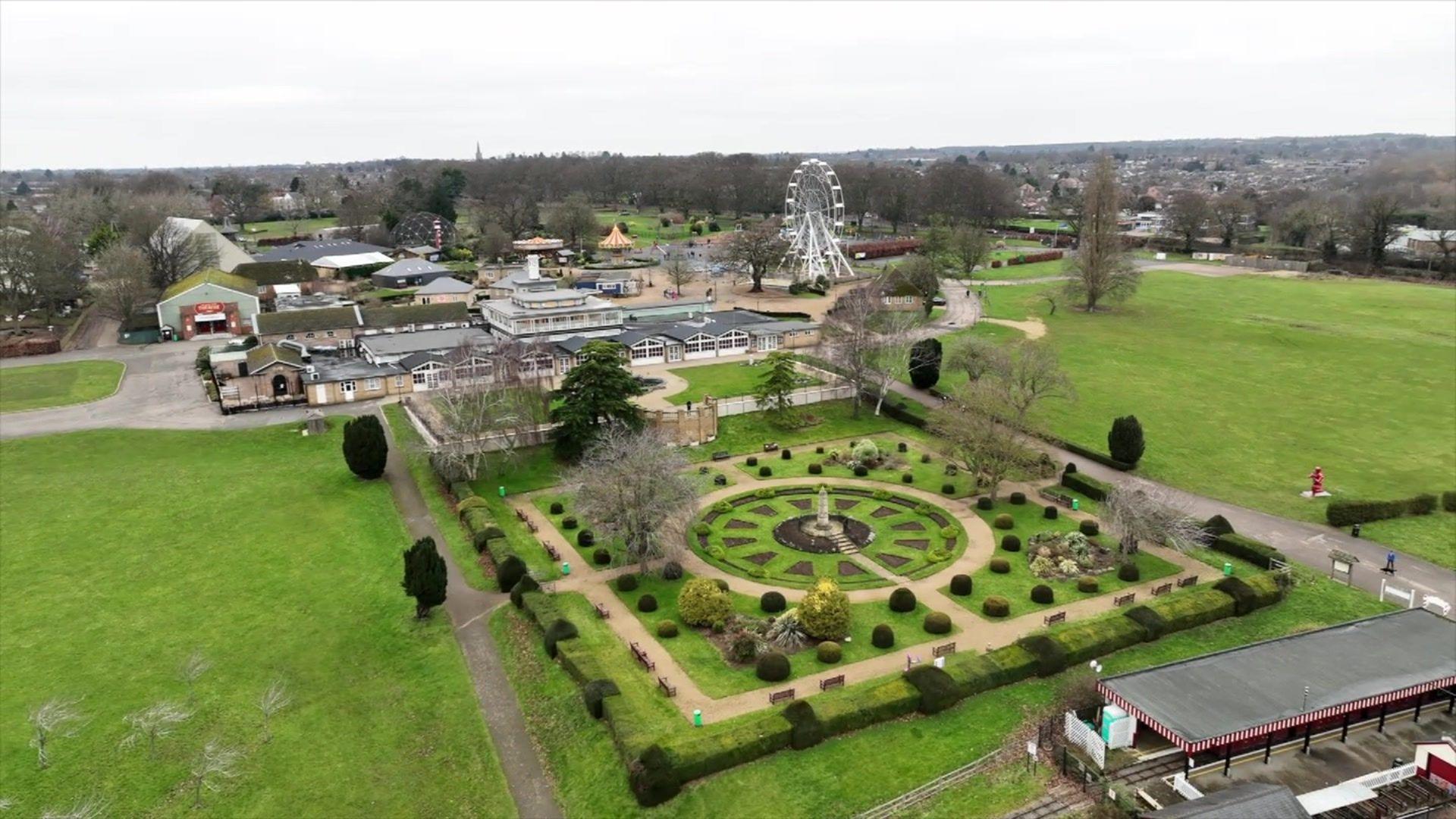 Drone shot of Wicksteed Park showing formal garden laid out as a central circle surrounded by squares with bushes laid in rows. A large pavilion building, other smaller buildings and a big wheel are visible in the background.