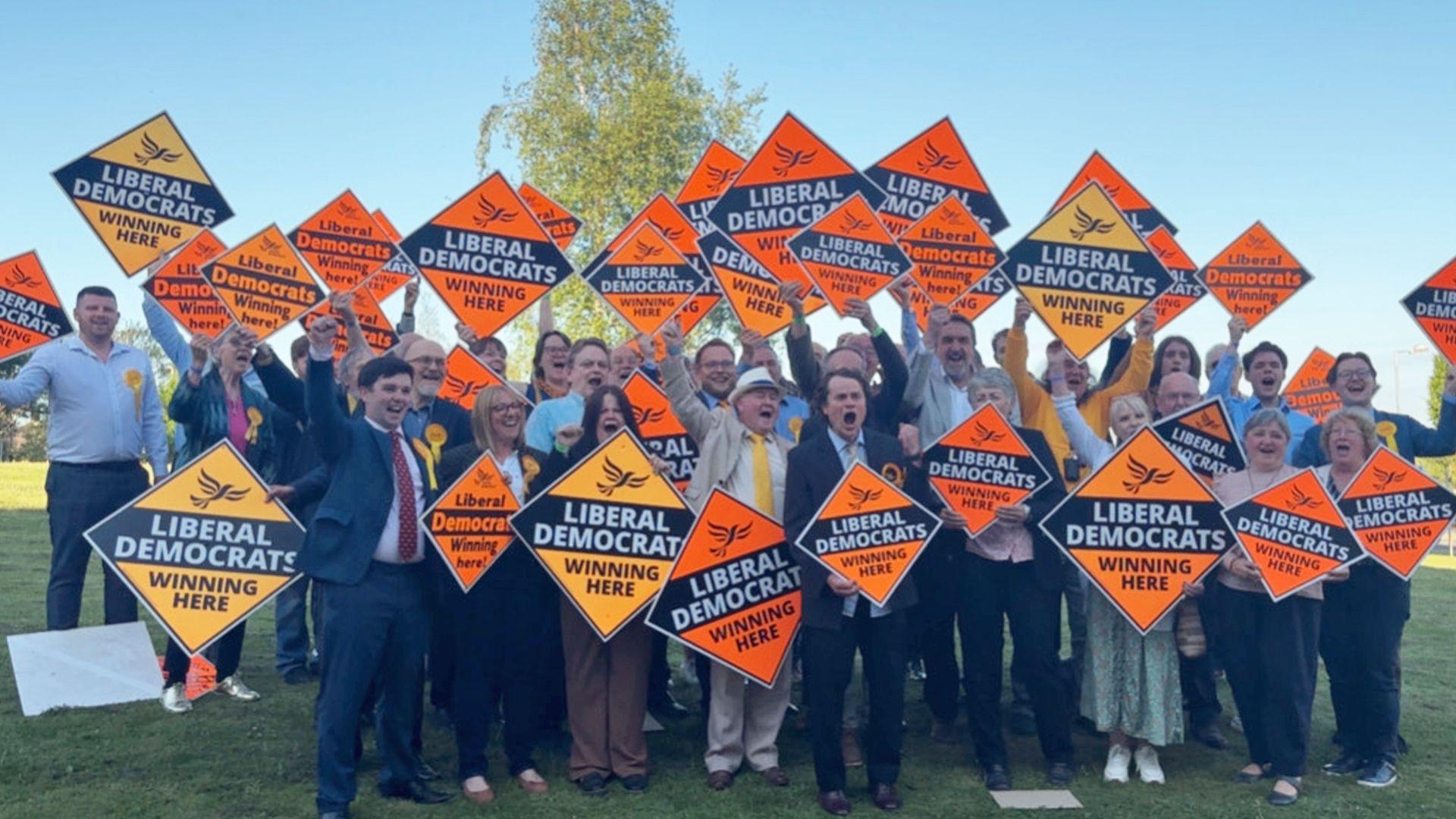 A group of jubilant campaigners stand on grass, holding up orange Liberal Democrat posters