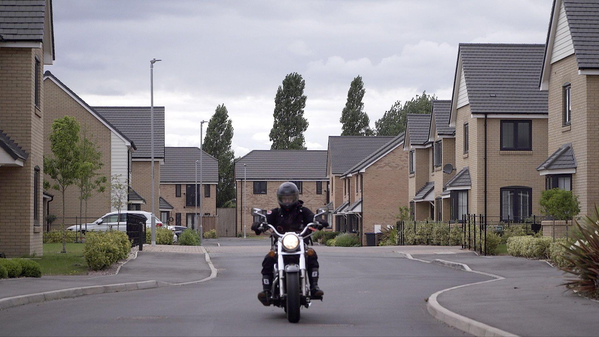 A rider on motorcycle heads towards the camera on an unmarked street in the Bassingbourn-Fields estate. New build houses with light brickwork and grey roofs line the street on either side and can be seen at the end of the road.