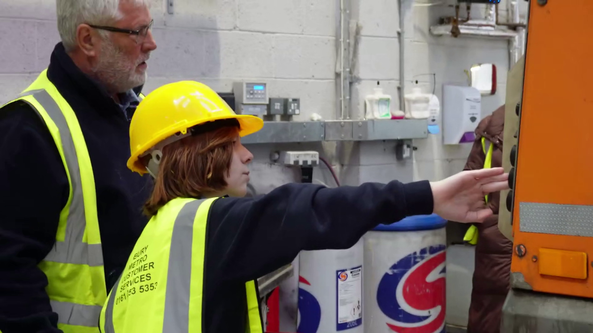 Tommy wears a yellow hard hat and high-vis vest as he presses a button on the back of a bin lorry to opperate it