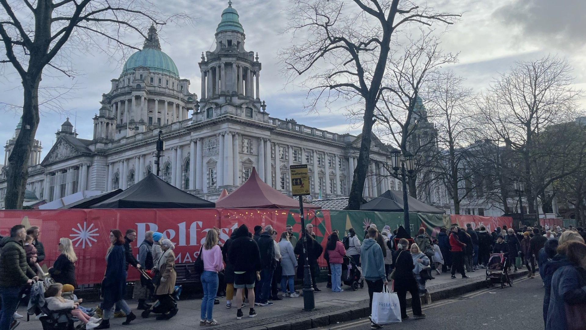 A queue of people stand outside Belfast's City Hall, there is a red and green banner across the gate with Belfast Christmas Market written in white writing. Belfast's white city hall is visible in the background.