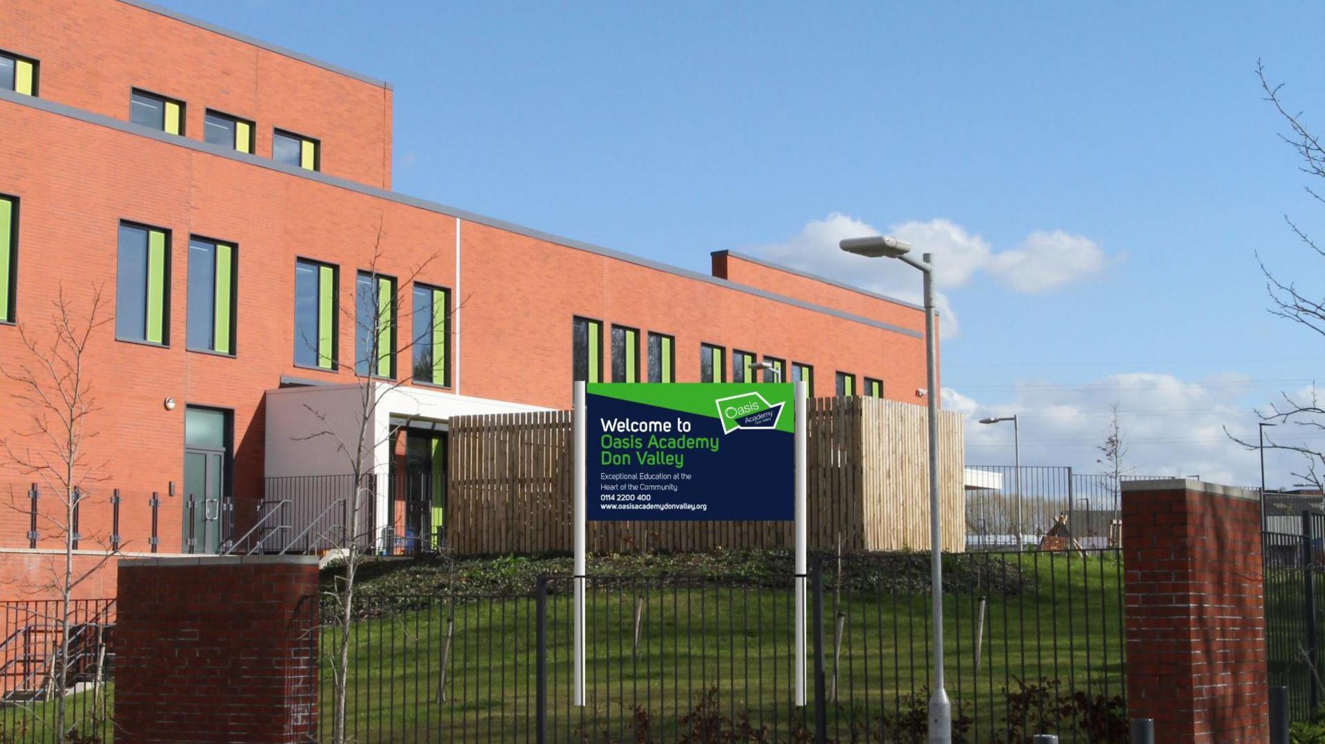 A modern building with a red-brick exterior and several rectangular windows featuring green frames. The structure appears to be multi-story. In front of the building, there is a fenced grassy area with some landscaping, including small shrubs and ornamental grasses. A sign is prominently displayed near the centre of the image, mounted on two poles. The sign reads:
“Welcome to Don Valley Academy” and includes additional text about the school and its trust affiliation.
The foreground consists of a paved walkway with several short, cylindrical bollards arranged in a line, likely to prevent vehicle access. There are also two brick pillars integrated into the fence. A tall streetlamp stands near the sign, and a few leafless trees are visible.