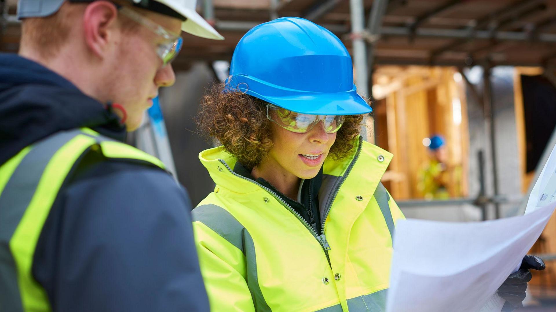 A man and woman wearing hardhats and hi-vis clothing looking at building plans on a building site