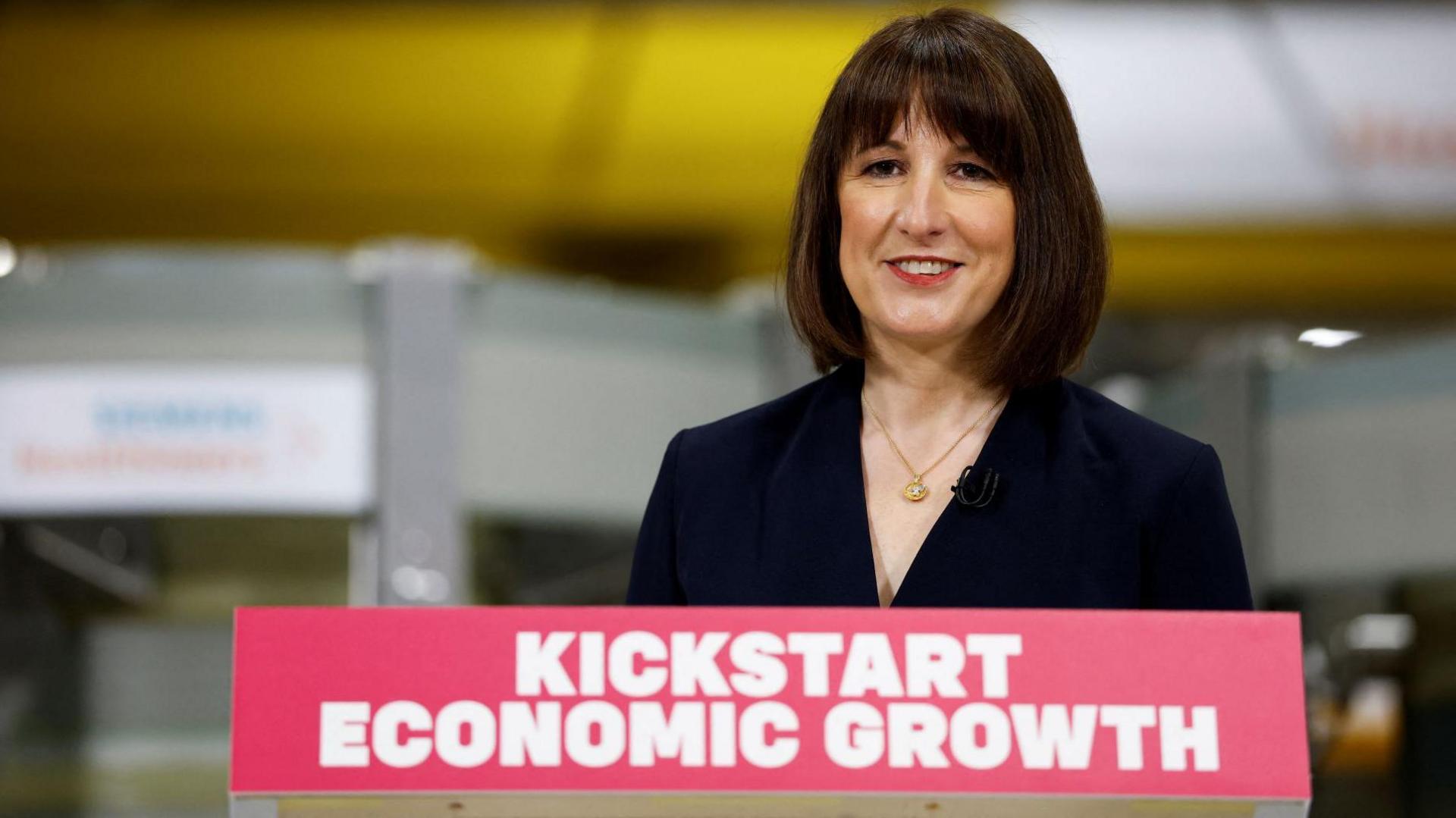 Rachel Reeves wearing a dark navy jacket stands behind a red sign saying kickstart economic growth in white letters.