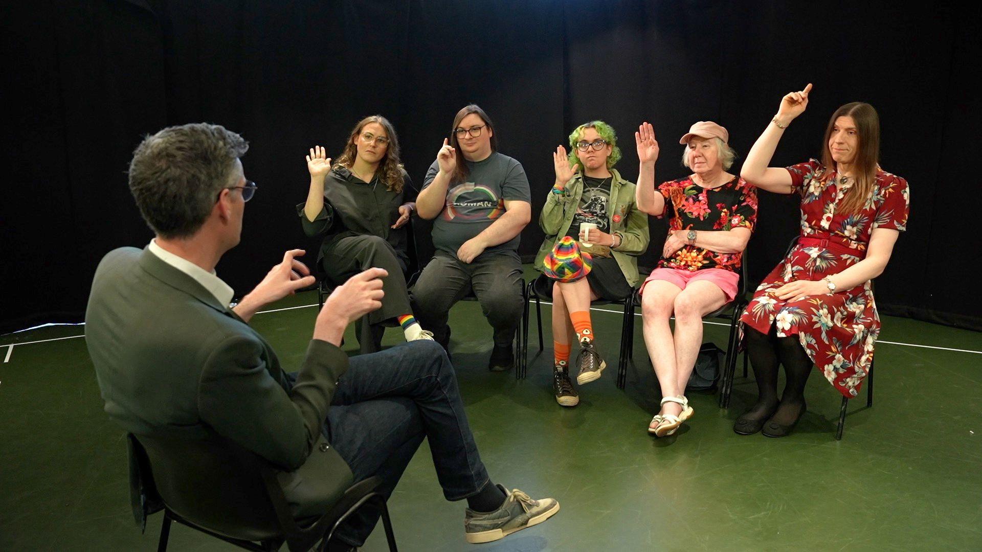 An interviewer with his back to the camera is faced by five people sitting in an arc on chairs. They all have their hands in the air.